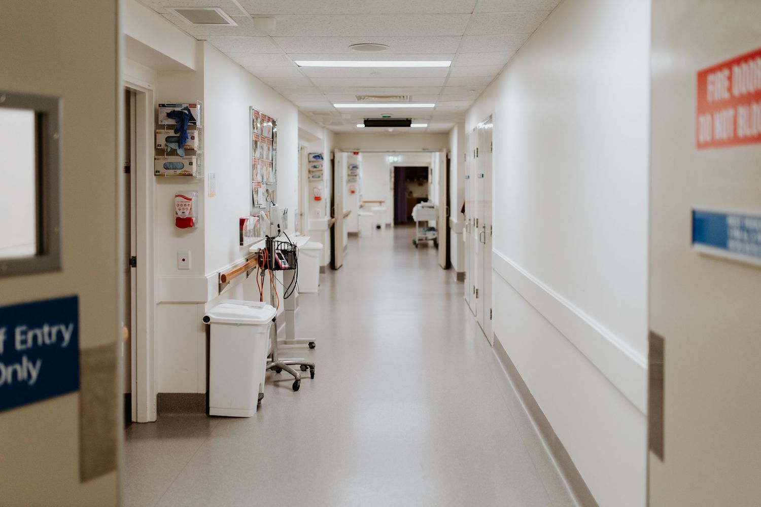 Empty hospital corridor with fluorescent lighting and white walls leads into the distance.
