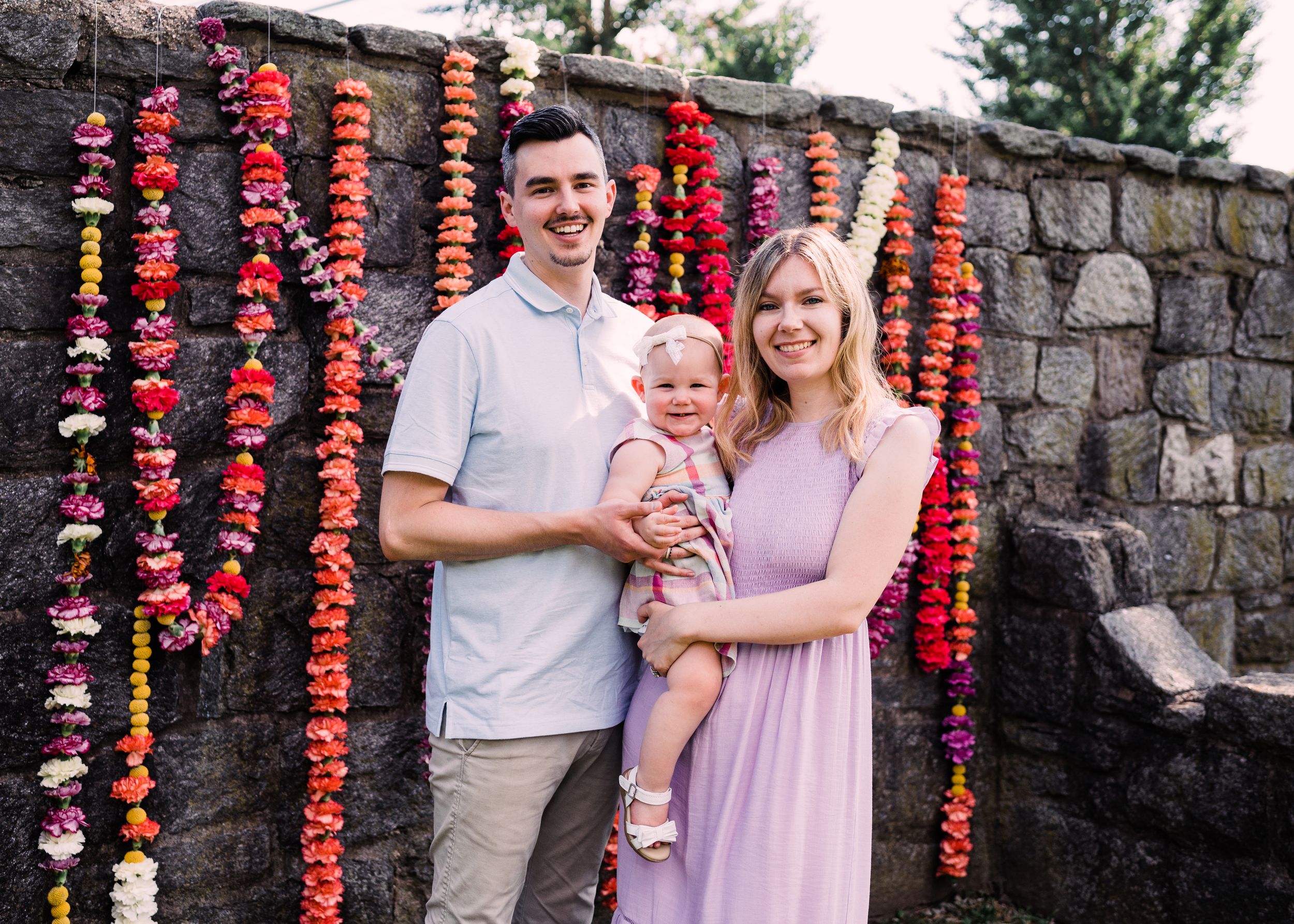 A family poses outdoors against a stone wall decorated with hanging strands of bright red paper flowers.