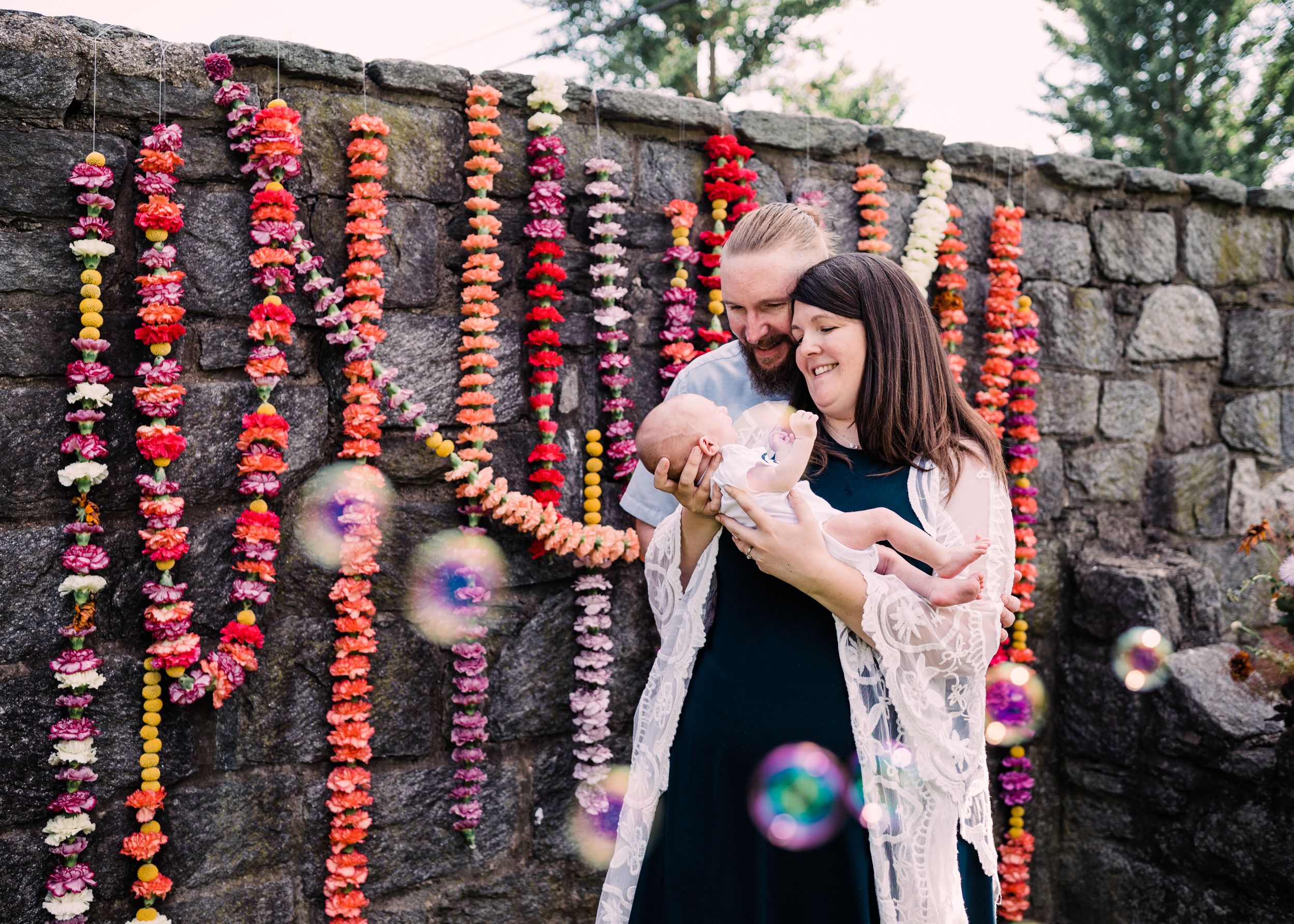Outdoor family moment captured with bubbles and colorful hanging flower garlands against a stone wall.