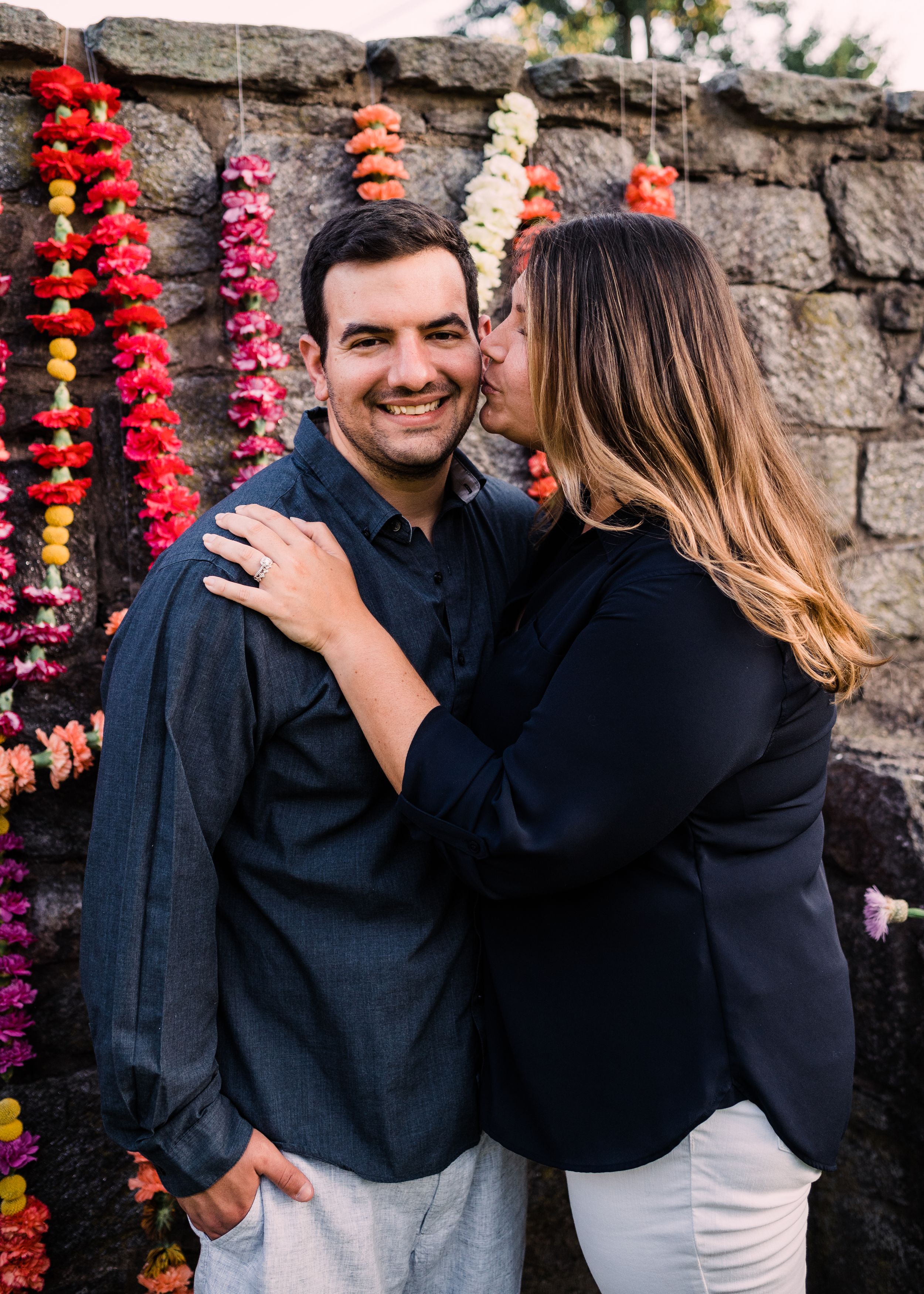 Couple in coordinated black outfits sharing intimate moments against a stone wall decorated with colorful hanging flowers.