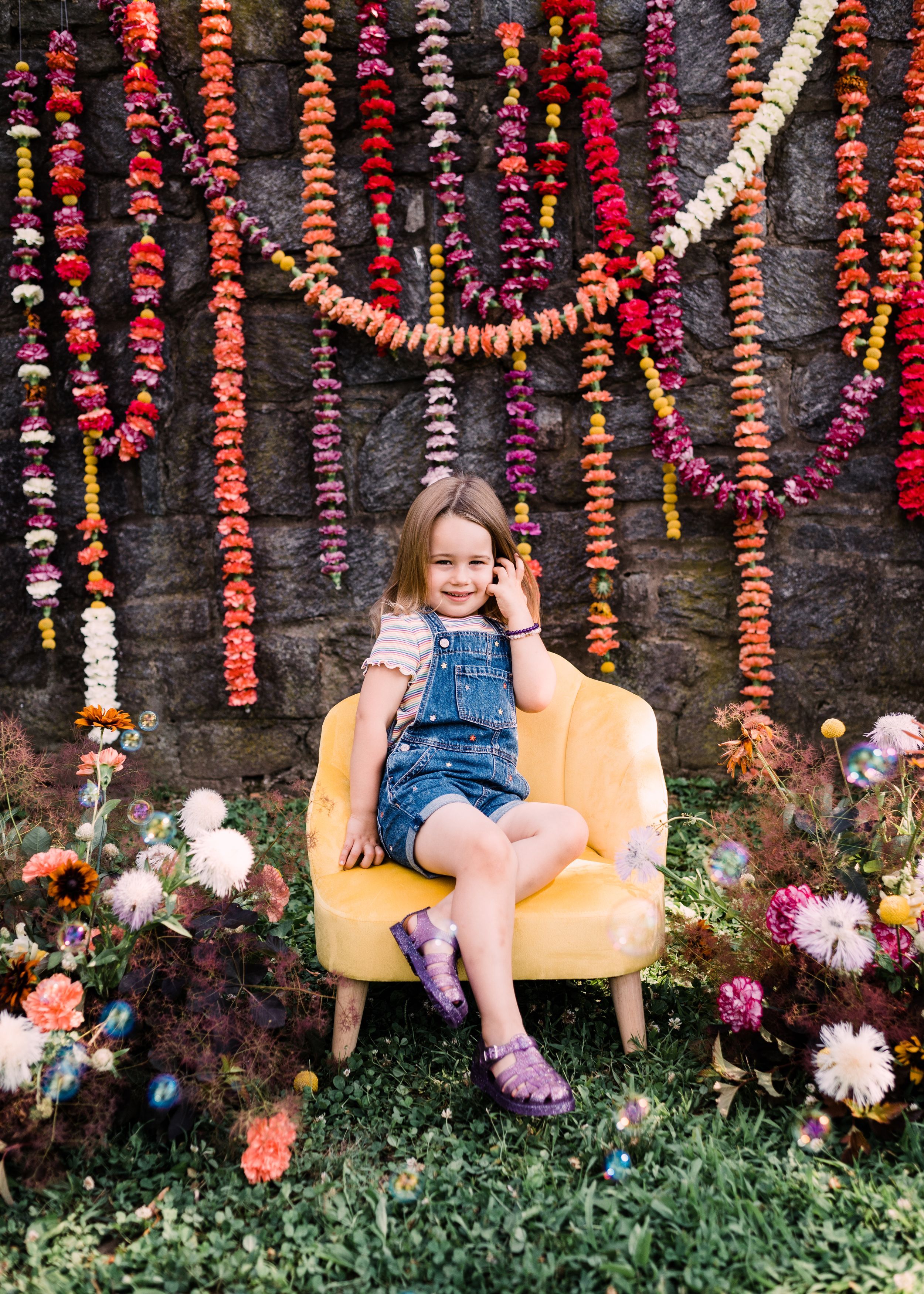 Yellow chair placed against stone wall with hanging colorful flower garlands and greenery below.