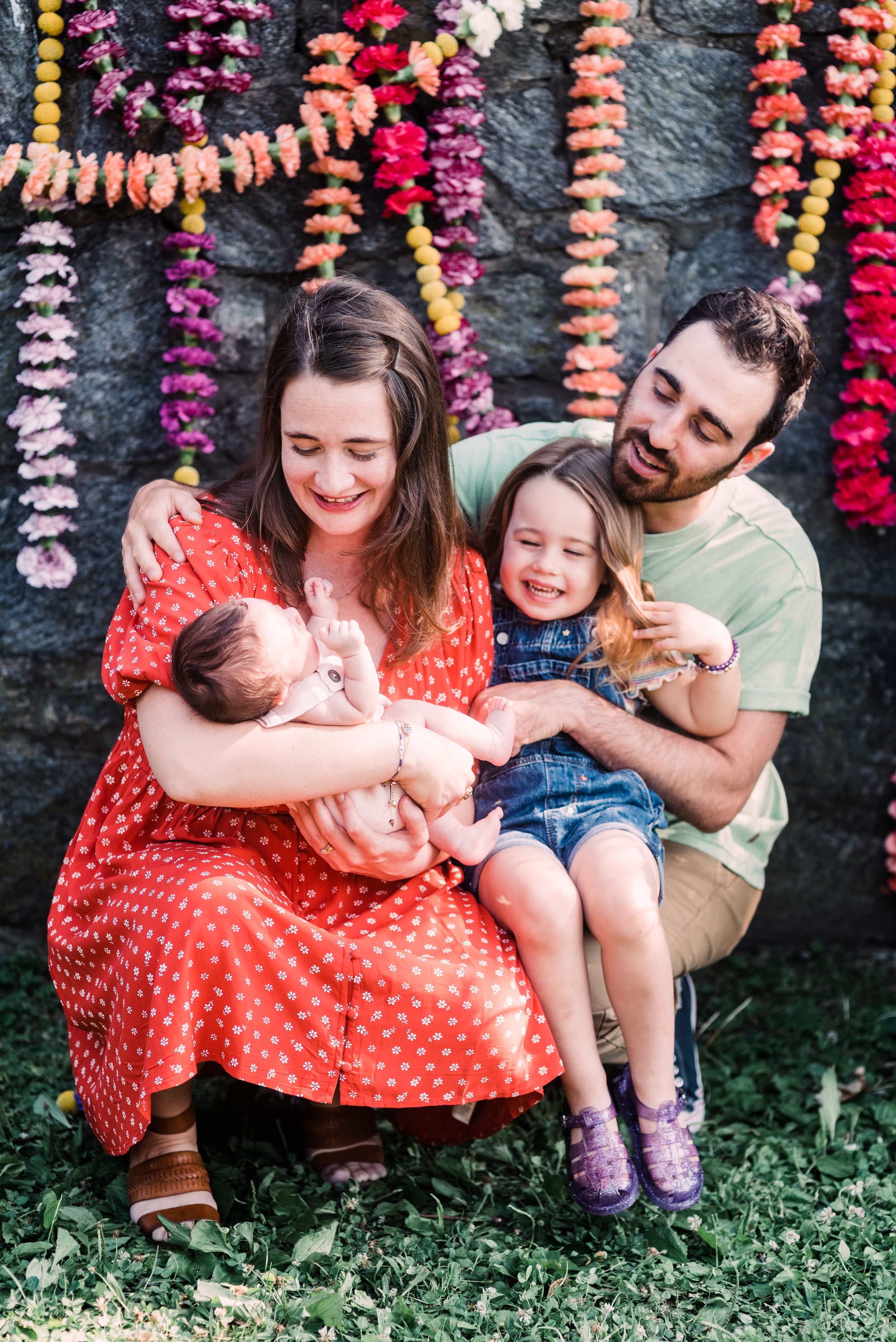 Family of three sitting and laughing together against stone wall with hanging colorful flower garlands.