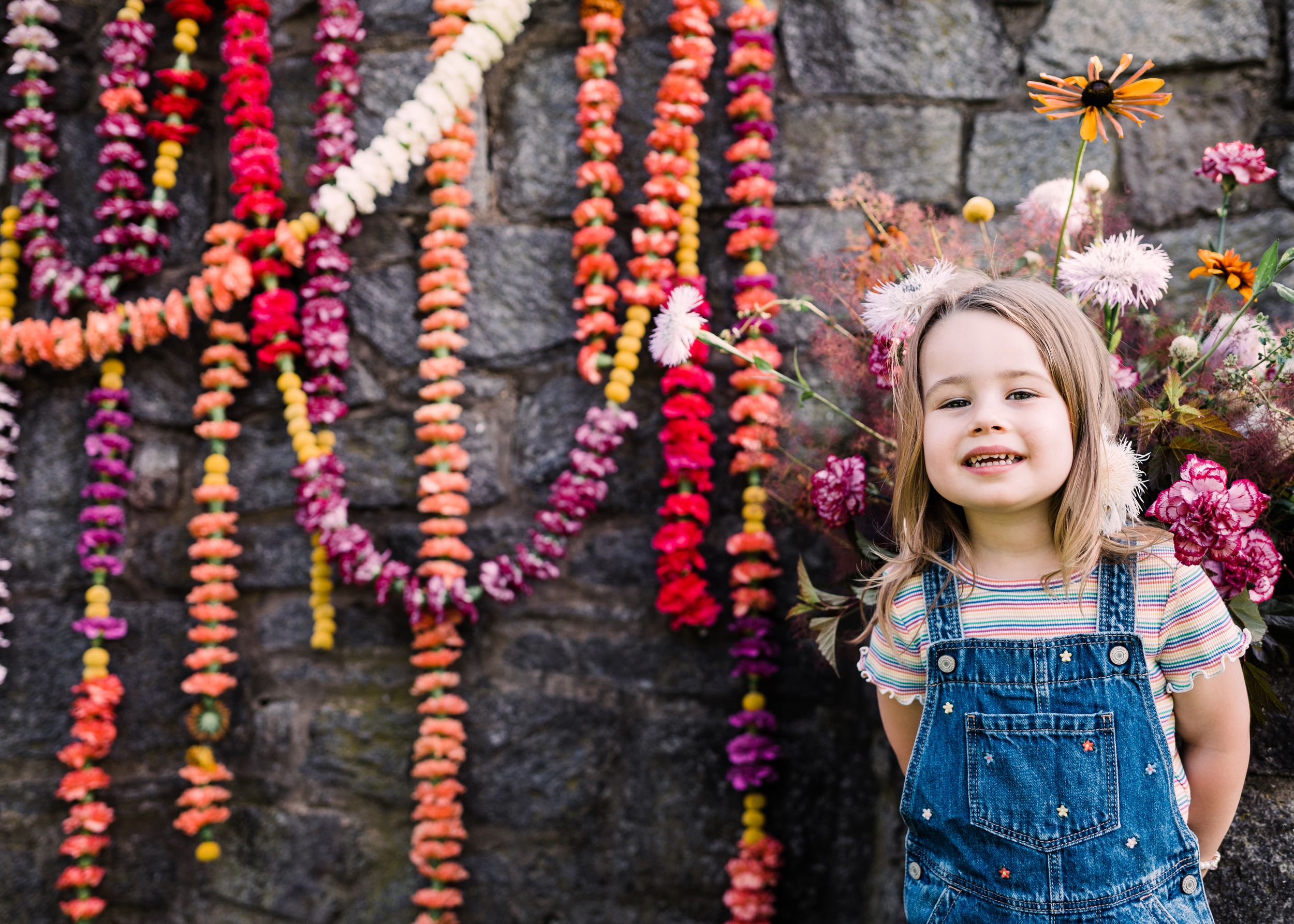 Child wearing denim overalls poses playfully in front of stone wall decorated with pink and red flowers.