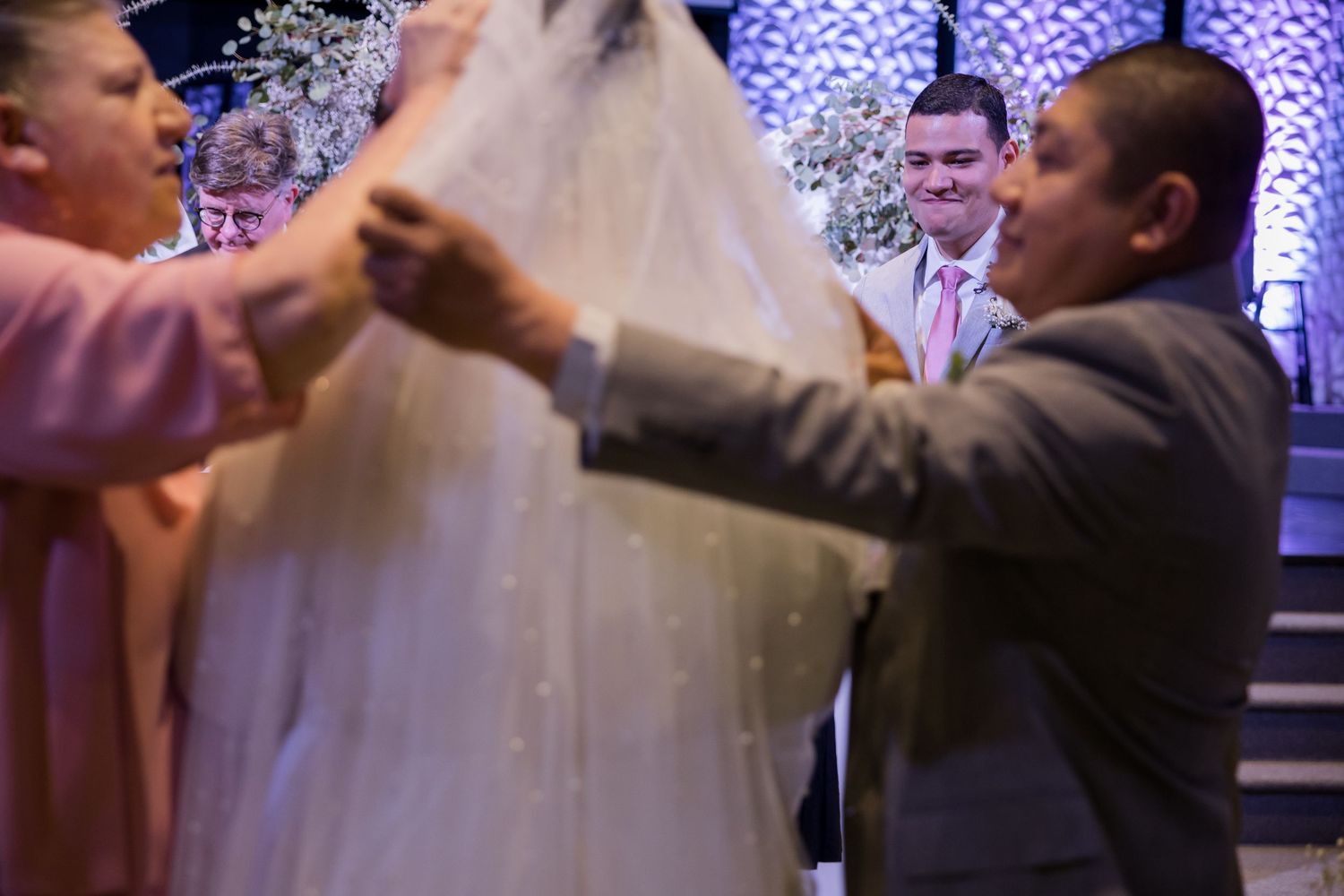 Two people help keep a bridal veil in place during a wedding celebration with purple lights in the background.