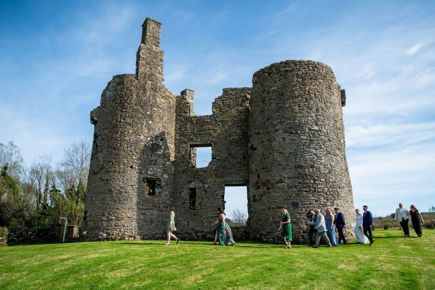 Ancient stone castle ruins against blue sky with visitors exploring the historic site on green grass.