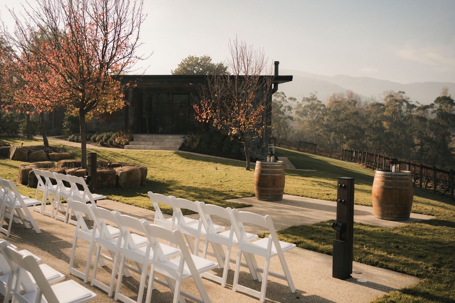 Row of white wedding chairs arranged outdoors with mountains and wine barrels in background.