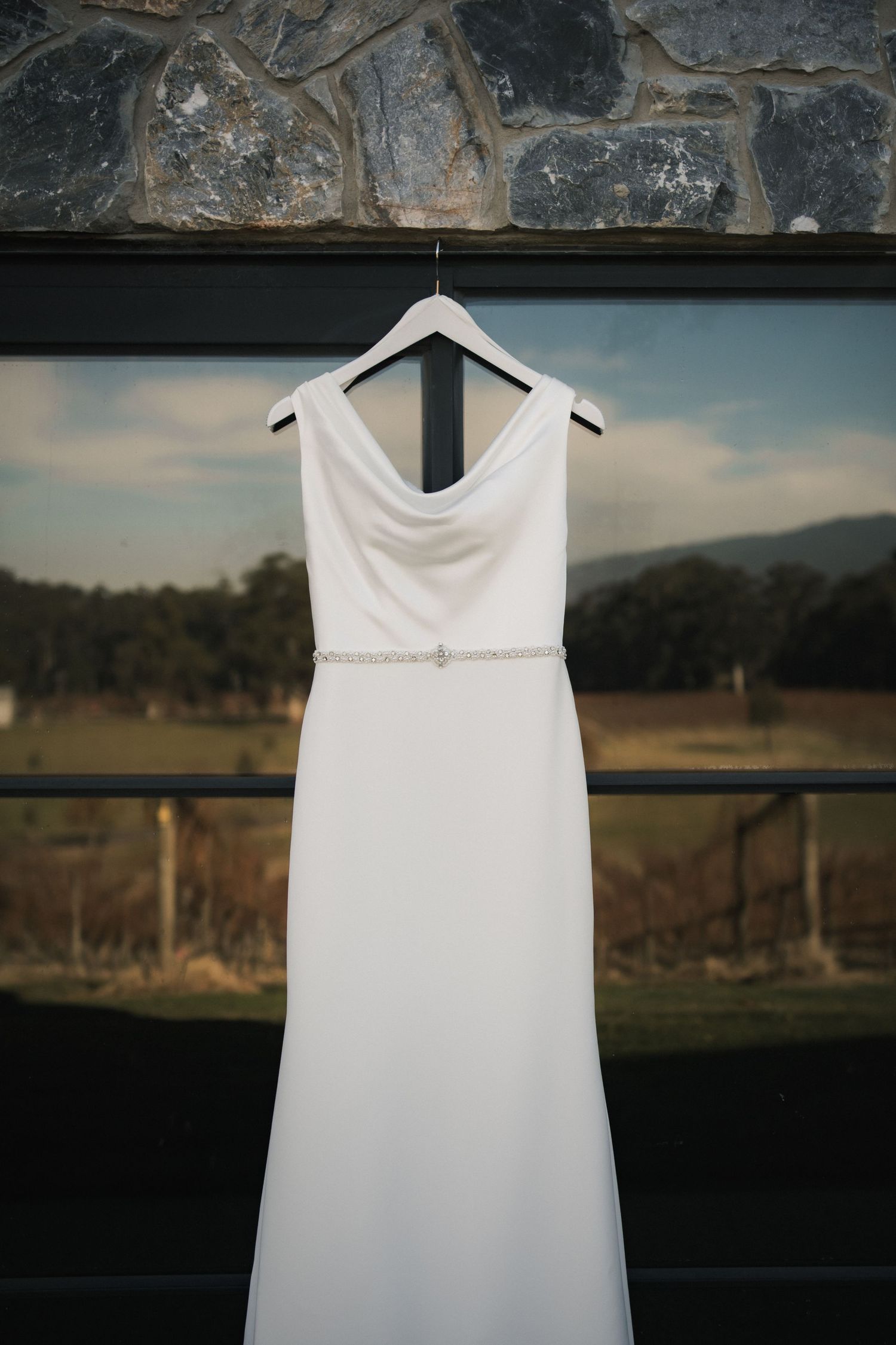 Elegant white wedding dress hangs on display against stone wall and scenic backdrop.