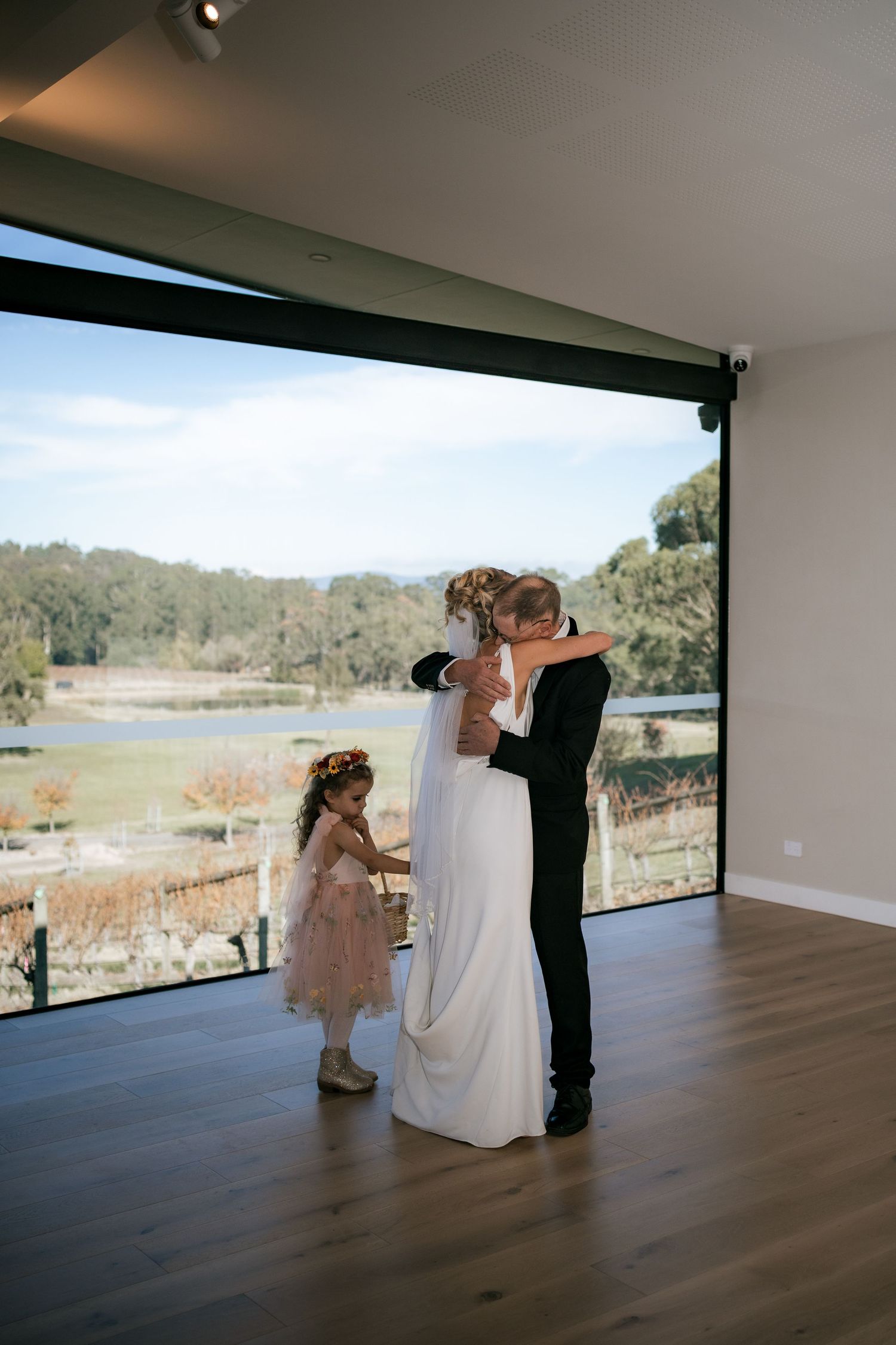 Wedding couple dancing in a modern venue with panoramic views while a flower girl watches.