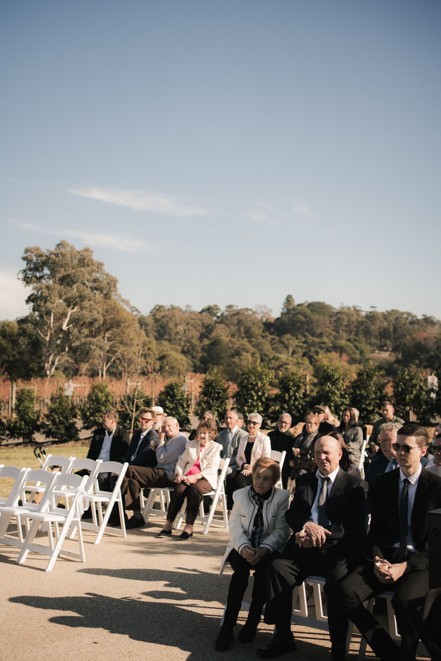 Wedding guests seated in white chairs at an outdoor ceremony venue with trees and blue sky in the background.