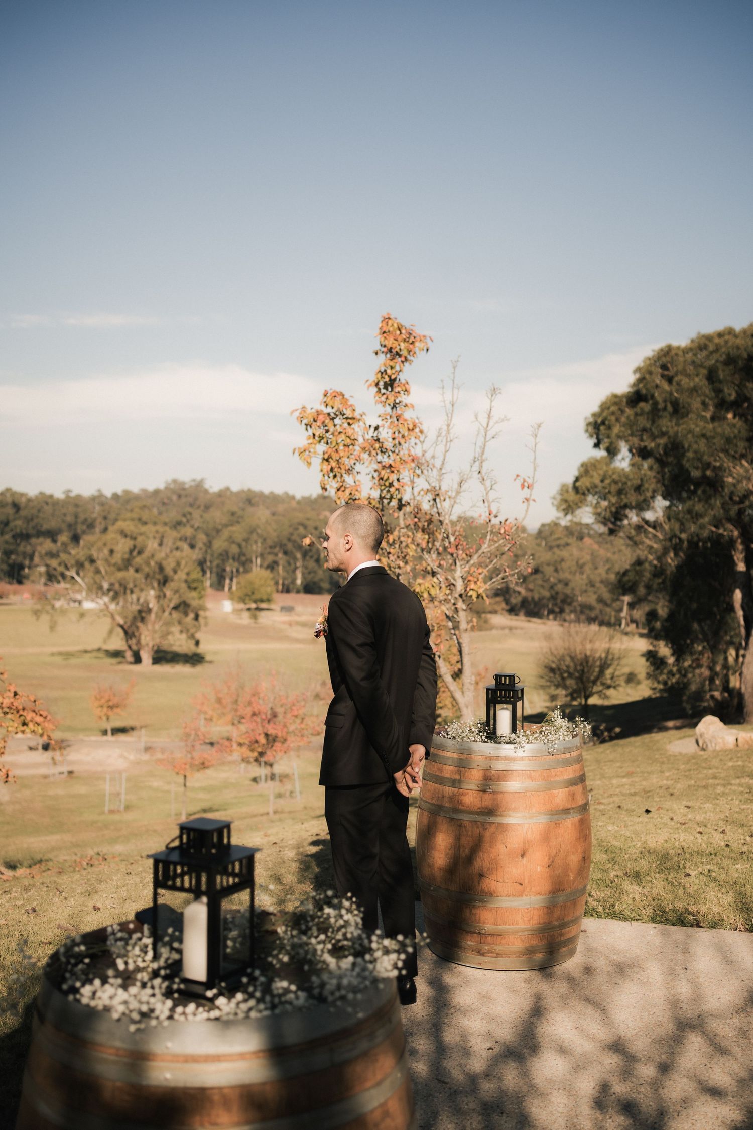 A rustic outdoor setting with wine barrels and lanterns decorating a grassy hillside overlooking a scenic landscape.