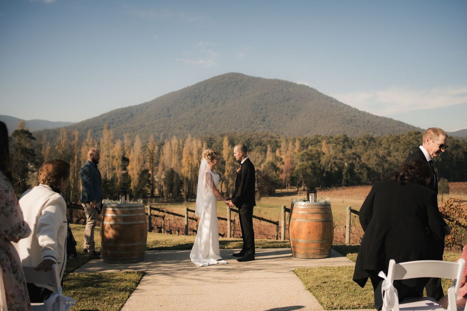 Outdoor wedding ceremony set against a mountain backdrop with wine barrels and golden trees at sunset.