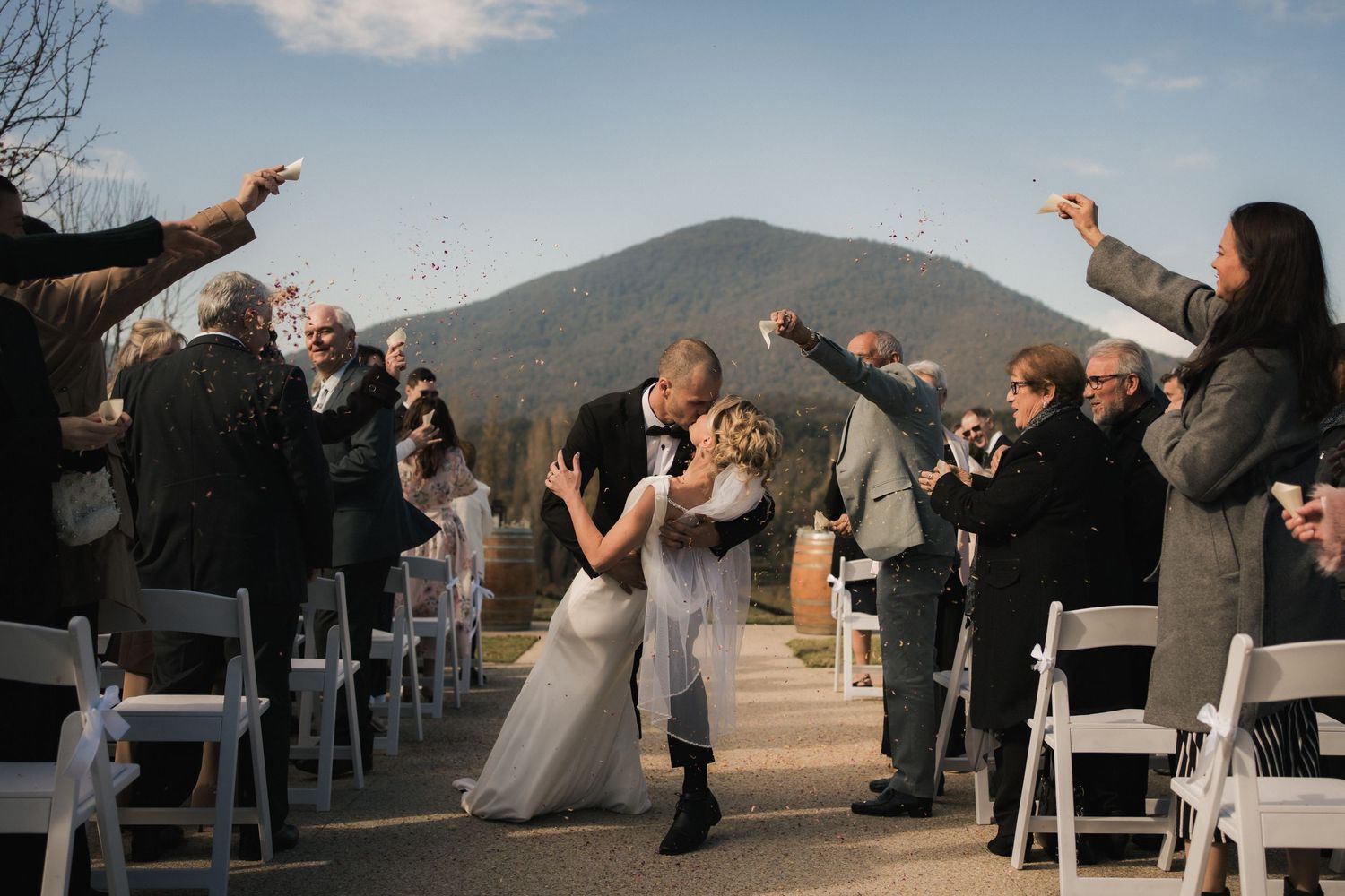 Wedding guests cheer and raise their arms during an outdoor mountain ceremony with white chairs and scenic views.