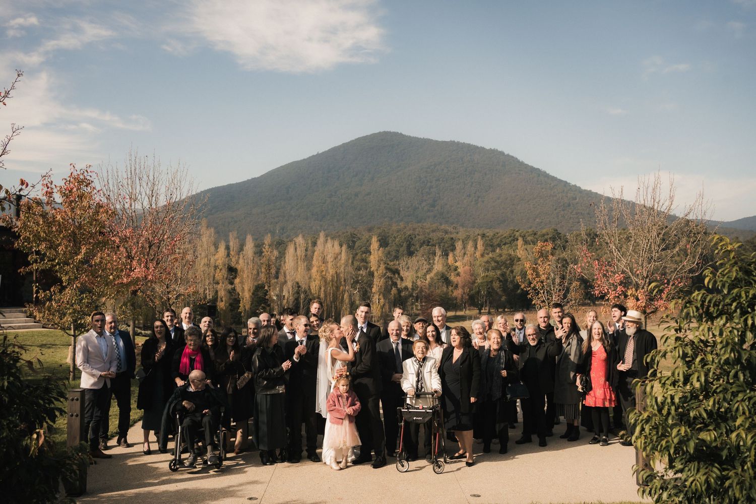 Wedding party wearing formal attire gathered in front of mountains with autumn trees in a panoramic outdoor setting.