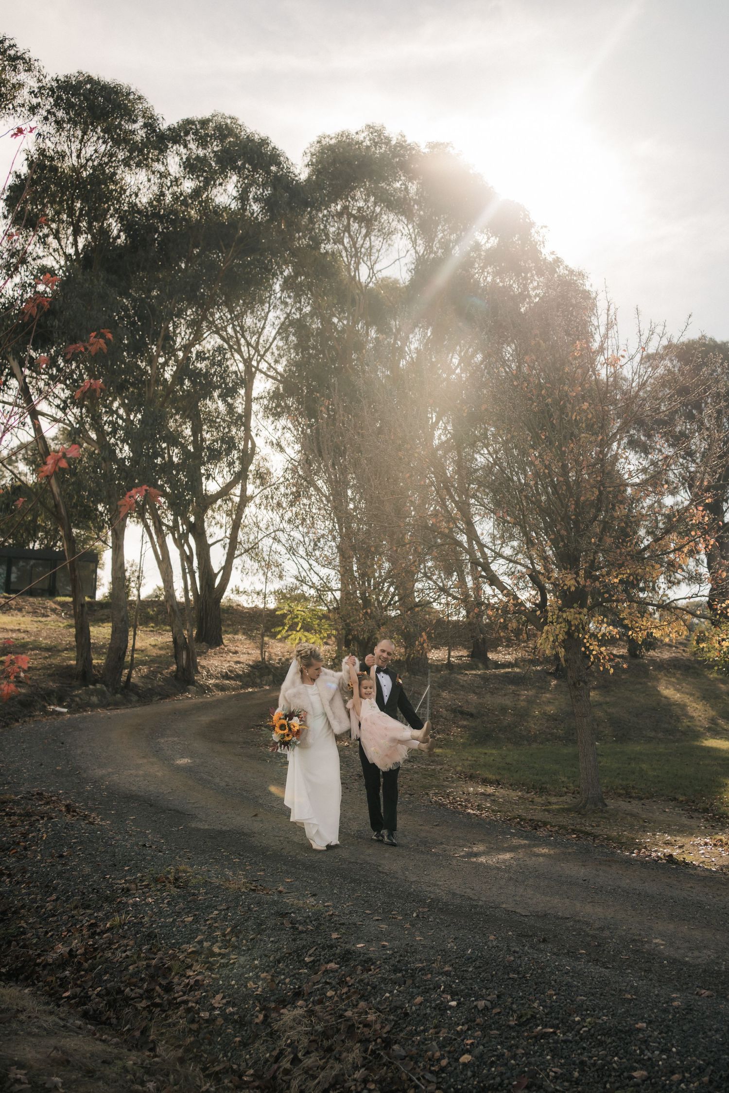 Newlyweds walking down a dirt path at sunset with eucalyptus trees silhouetted against bright sky.