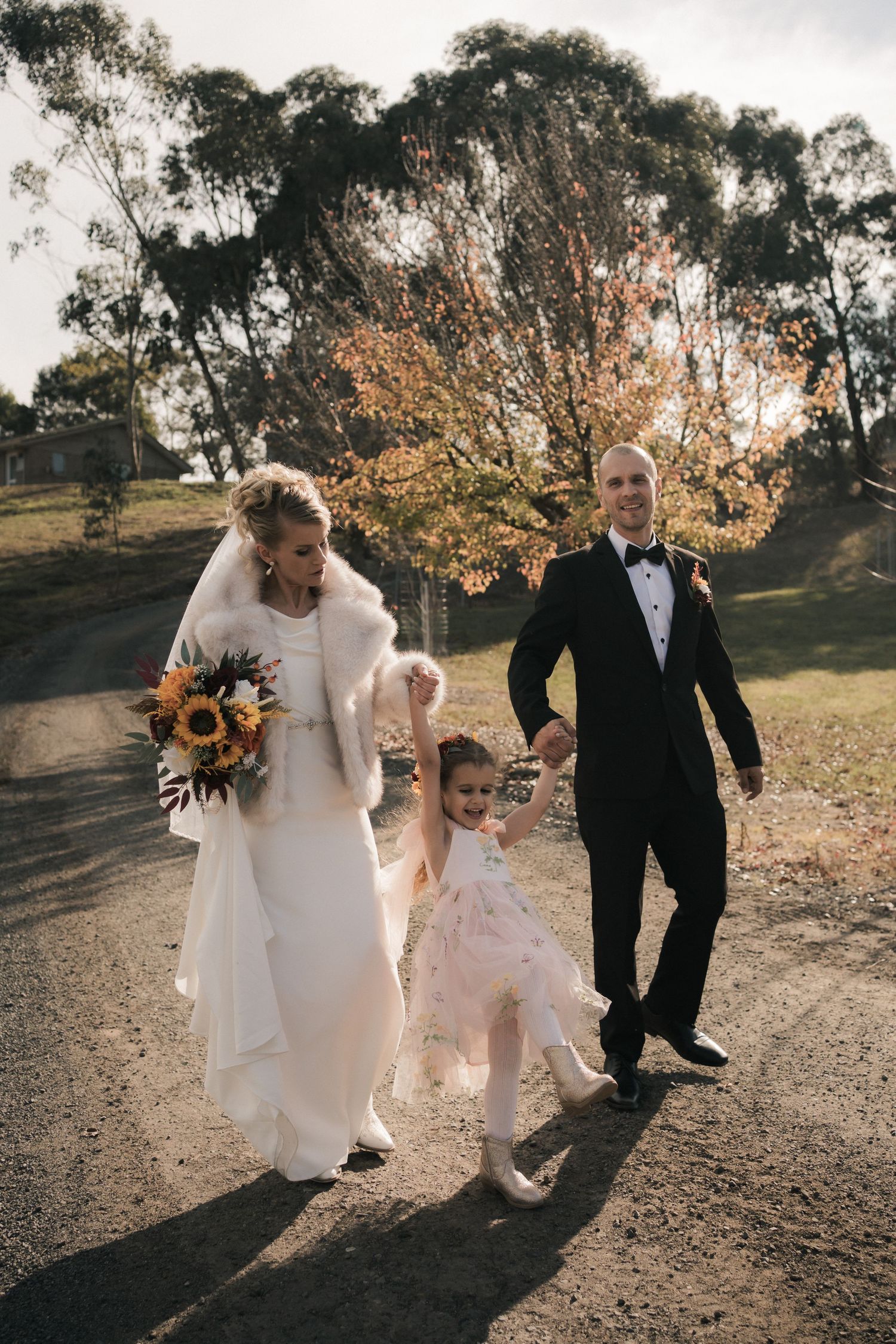 Wedding couple walks down path with flower girl while carrying sunflower bouquet against autumn trees.