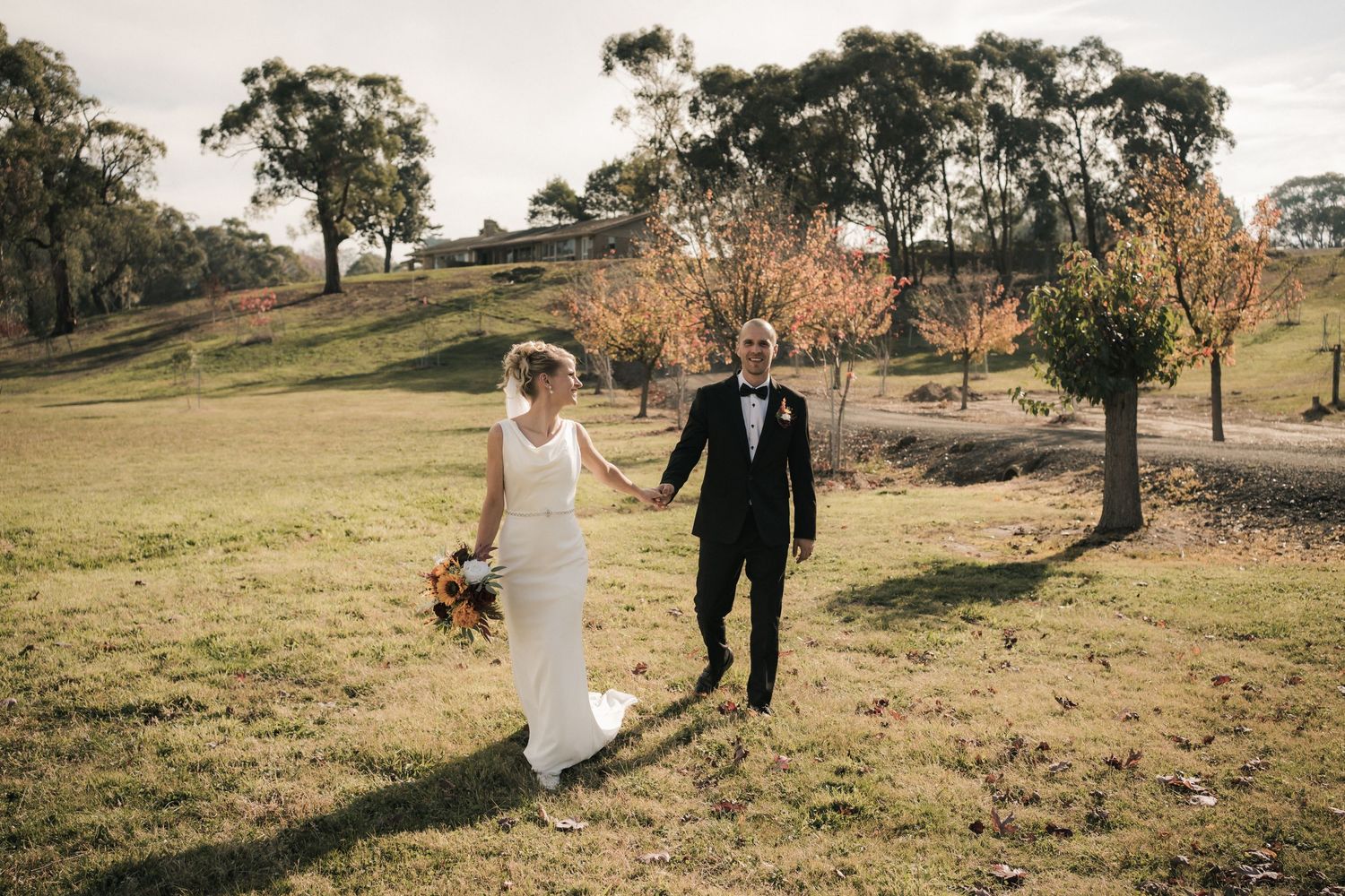 A couple dressed in wedding attire walks hand in hand through a scenic hillside with autumn trees in the golden sunlight.
