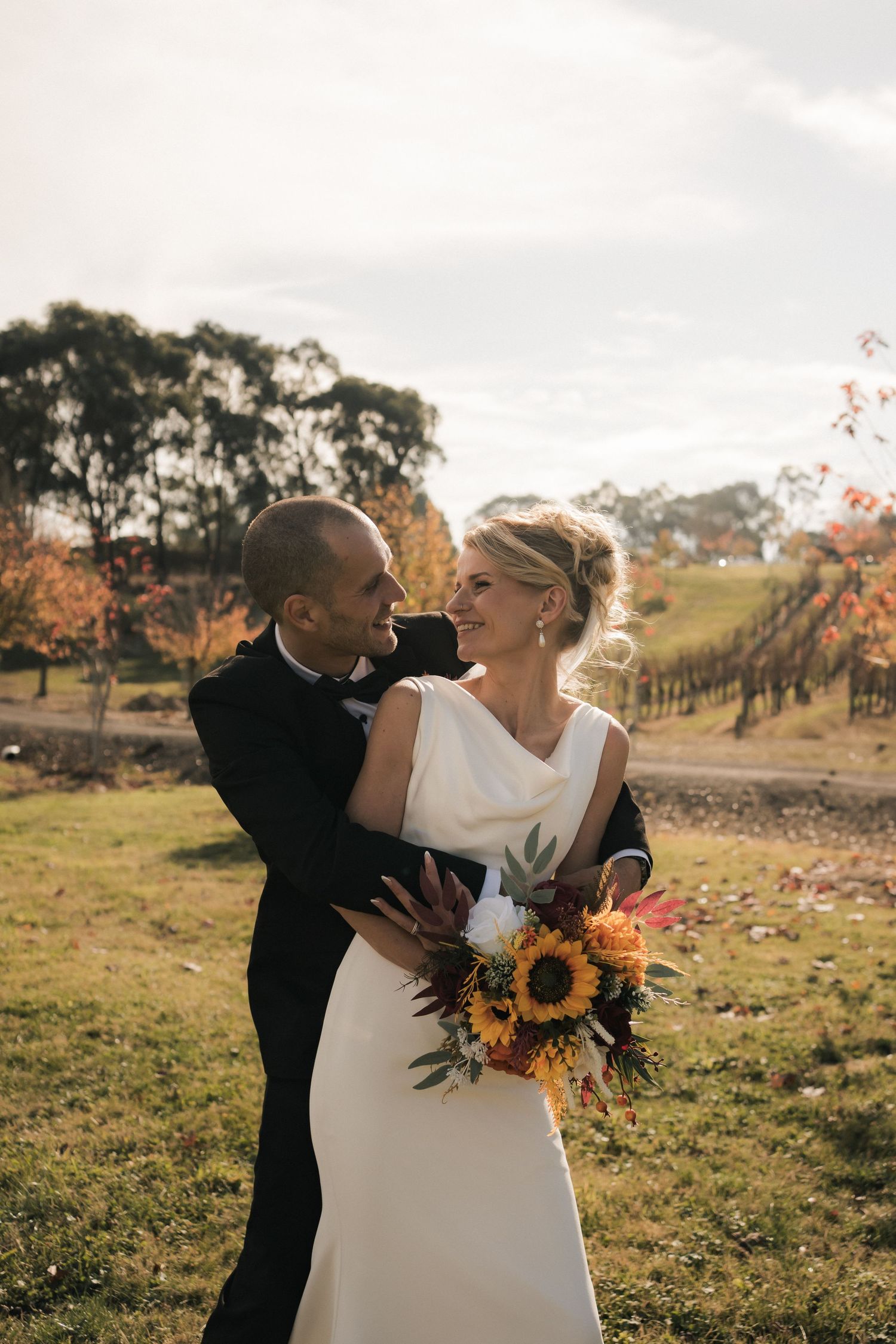Newlyweds share romantic moment in outdoor autumn setting with sunflower bouquet.