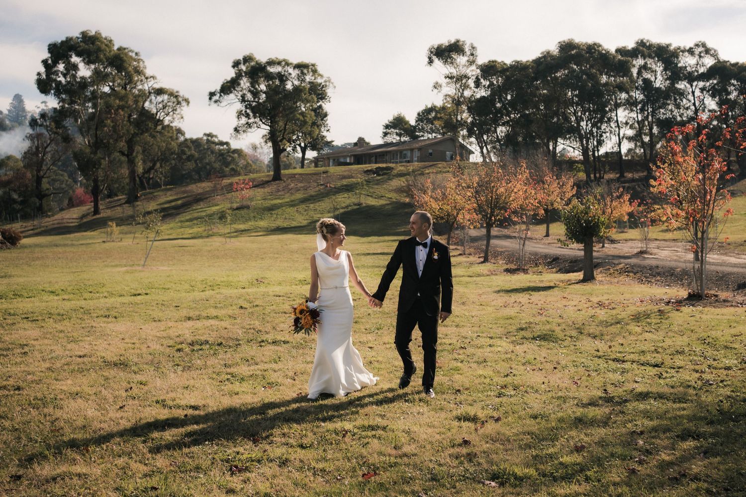 Two people in wedding attire walk hand in hand across a grassy hillside with tall eucalyptus trees in the background.