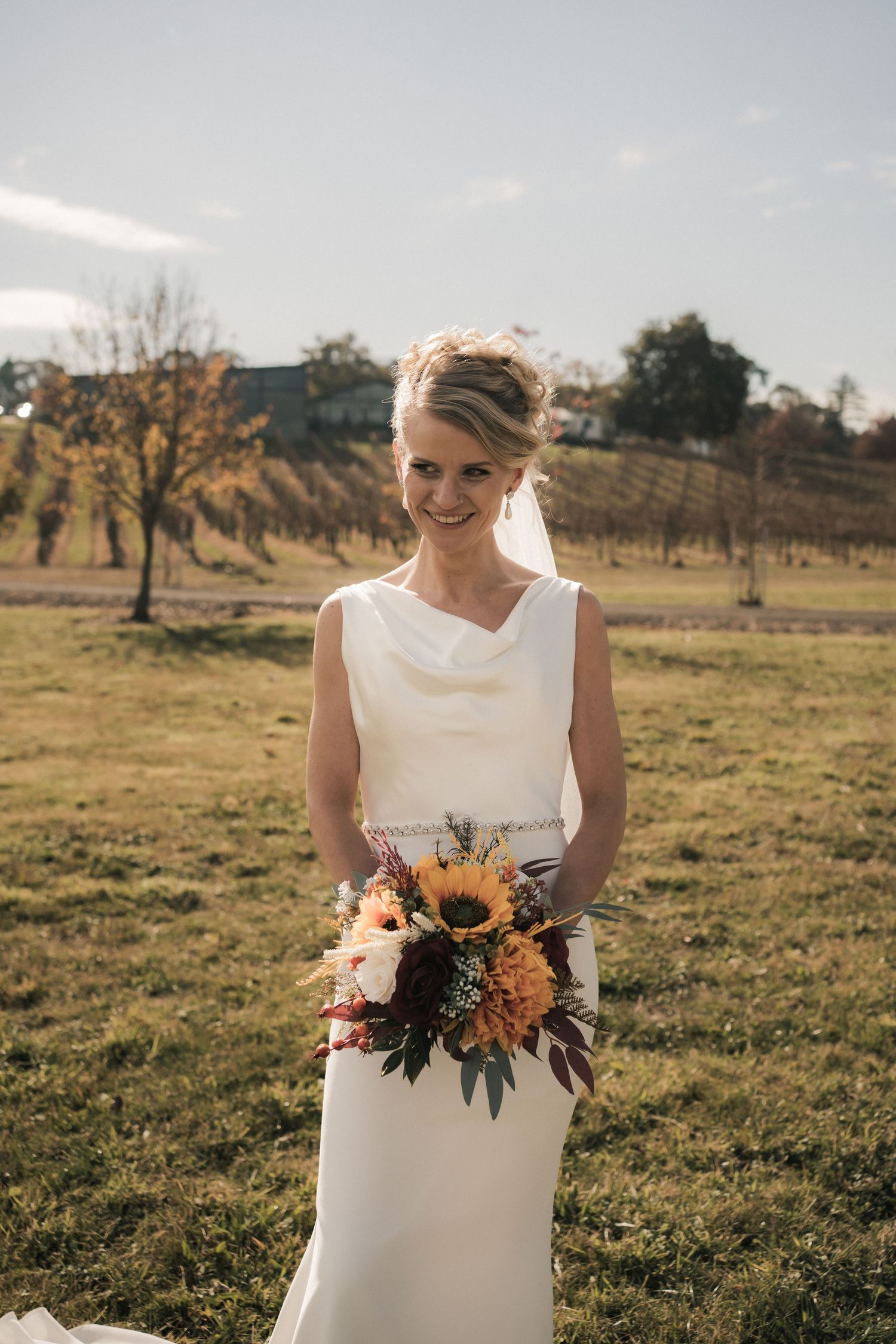 Bride in elegant white dress holds vibrant sunflower bouquet in rustic outdoor setting.