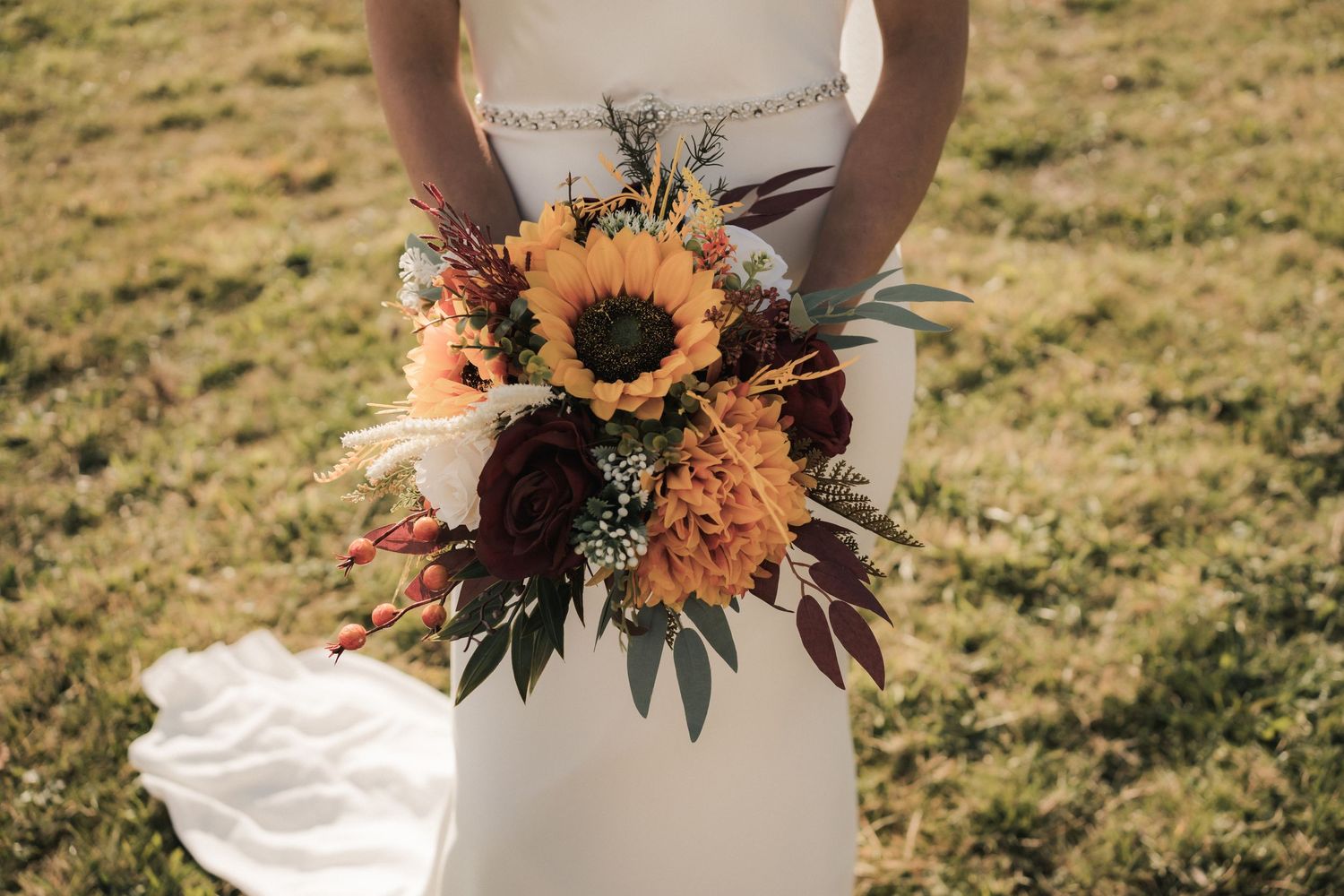 Wedding bouquet featuring sunflowers and autumn-colored flowers held against a white dress on a sunny field.