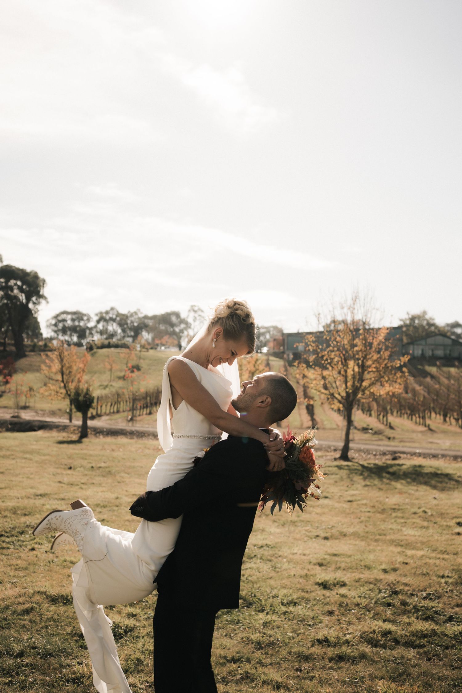 A romantic wedding couple embraces in an outdoor rural setting with rolling hills and scattered trees in the background.