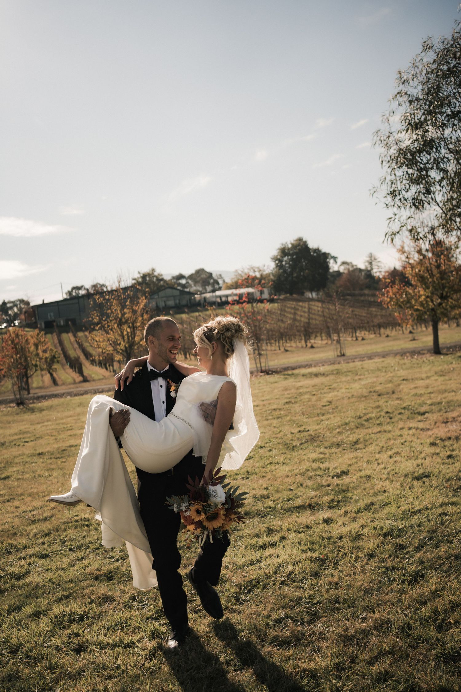 Wedding couple walking through a sunny field with golden grass and scattered trees.