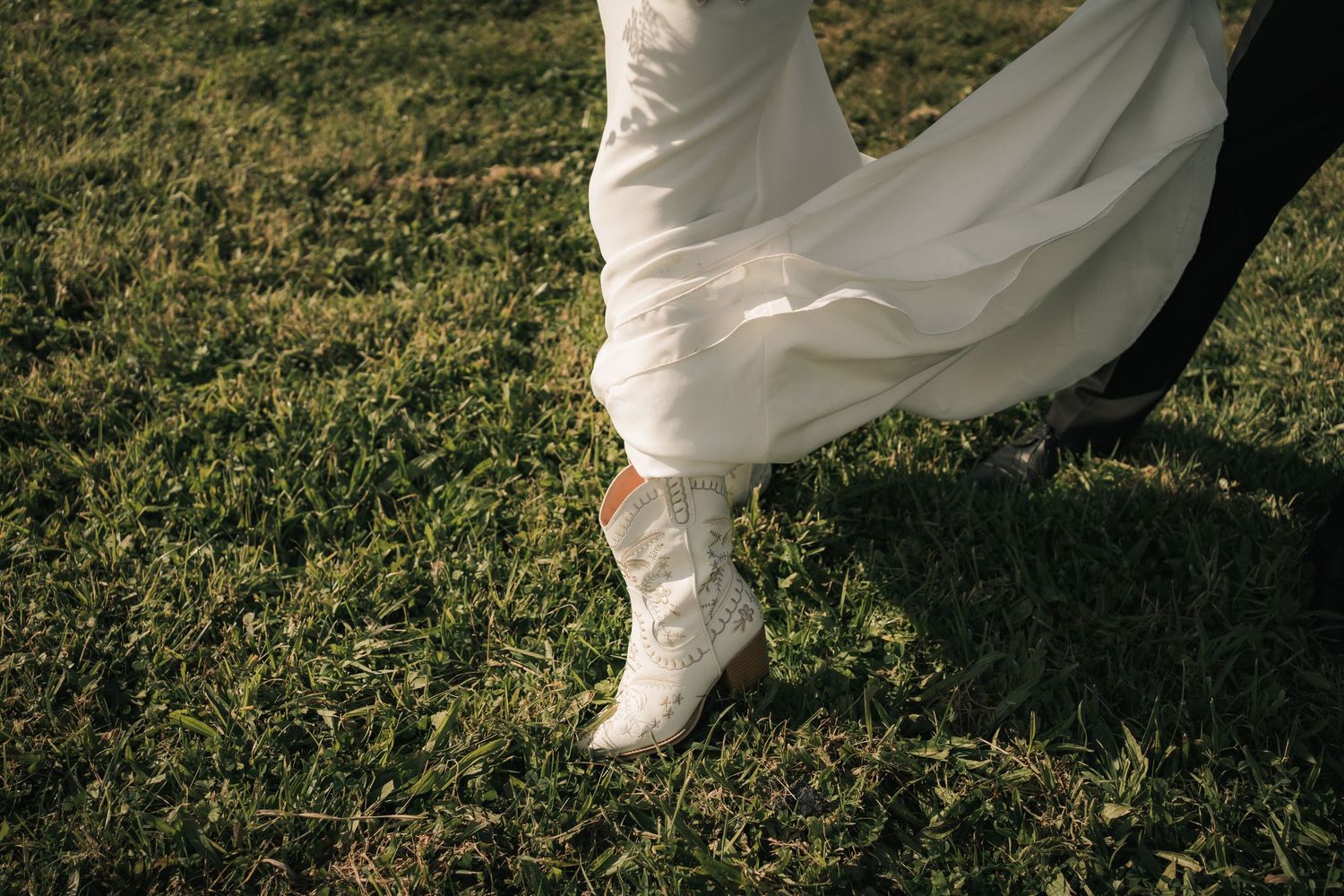 White wedding dress fabric flows elegantly over boots on green grass in an artistic detail shot.