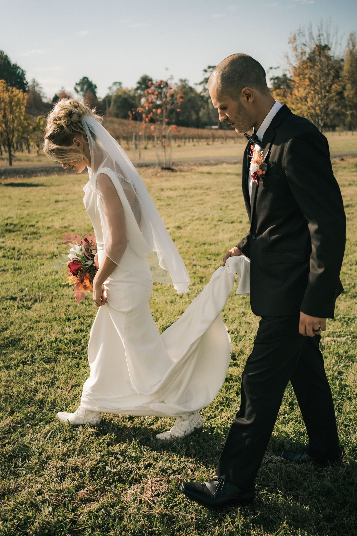 A groom walking behind his bride holding her dress train in an outdoor autumn setting.