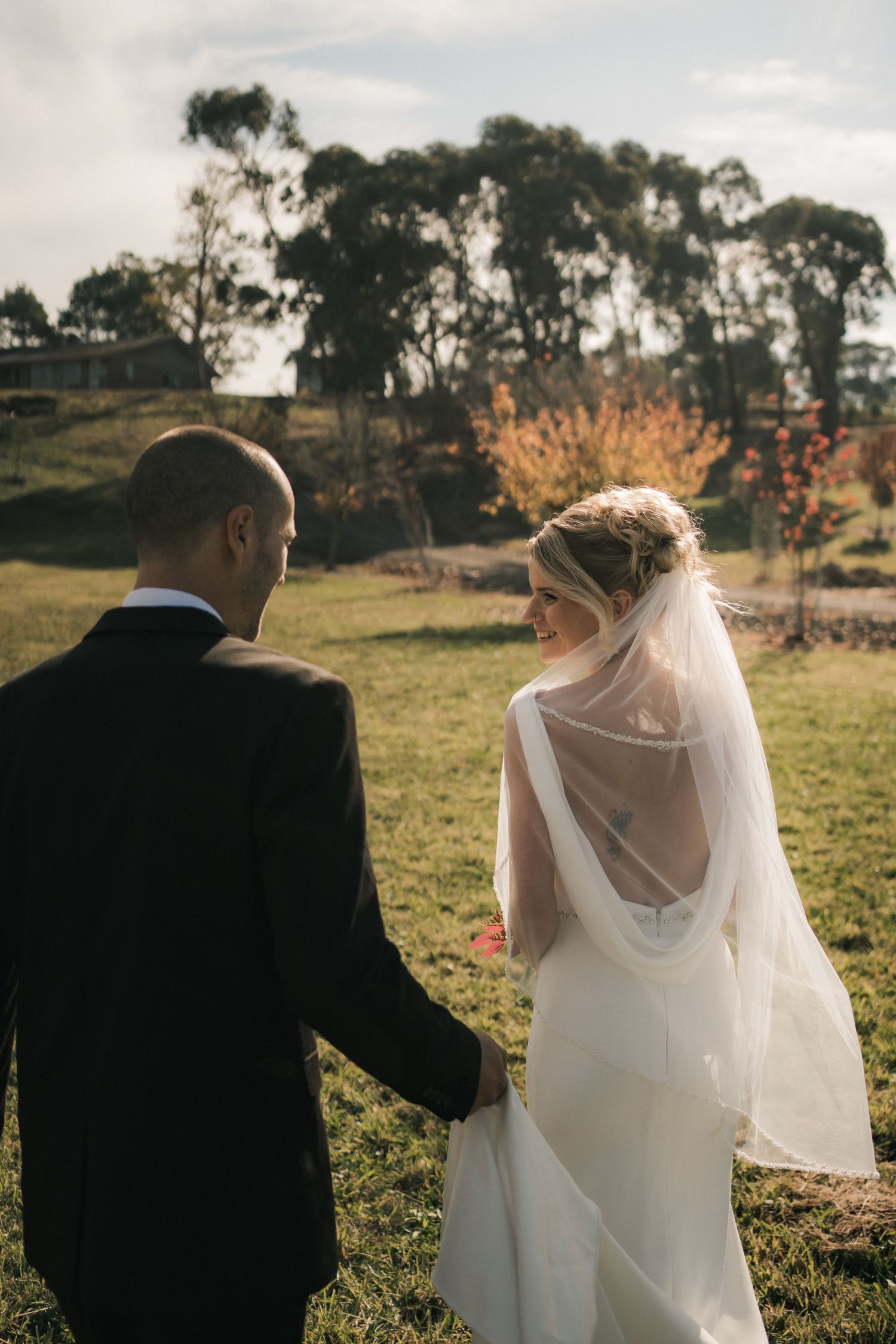 A back view of a couple in wedding attire walking on a grassy field with eucalyptus trees in the background at sunset.