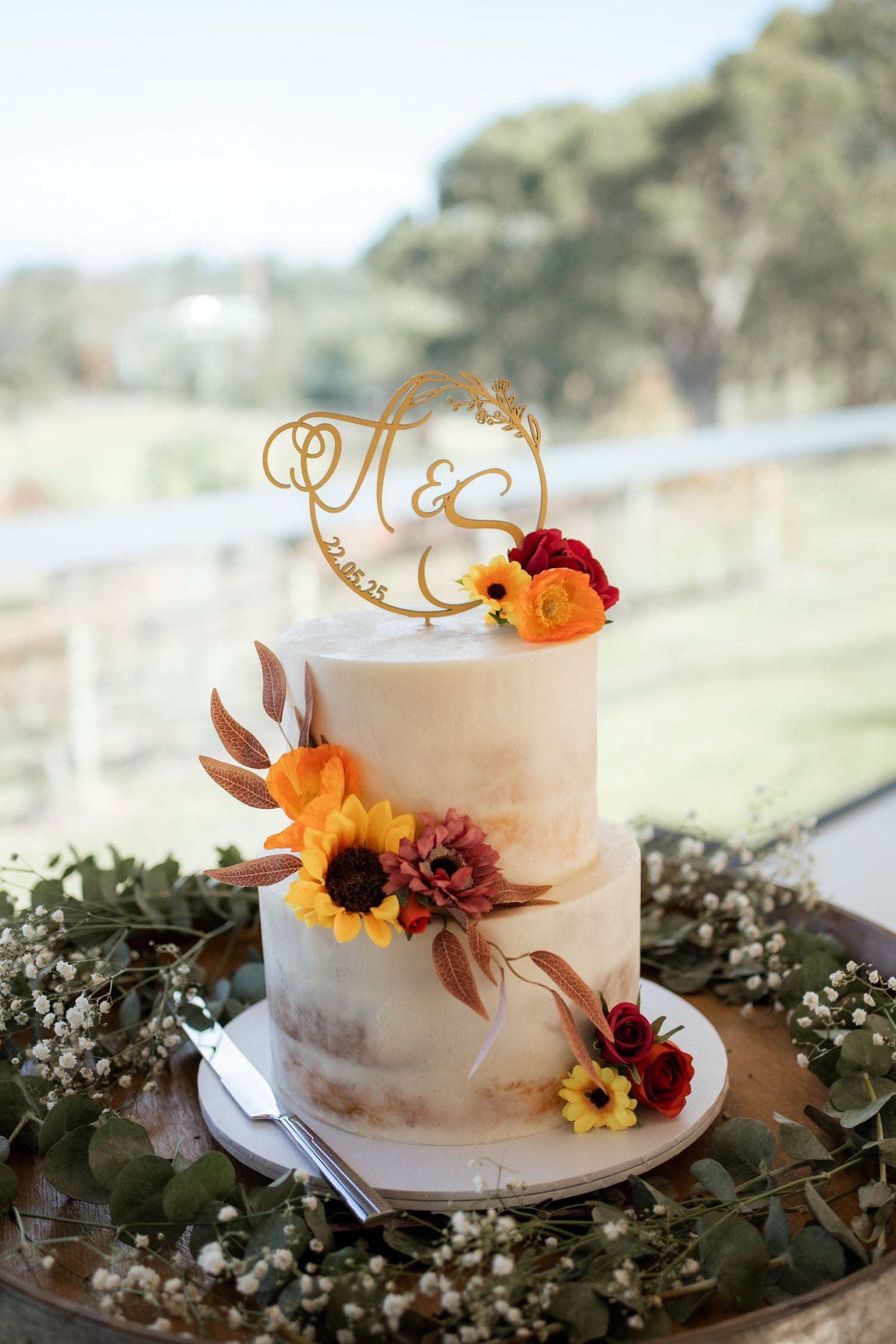 Two-tiered white wedding cake decorated with fall flowers and gold monogram topper.