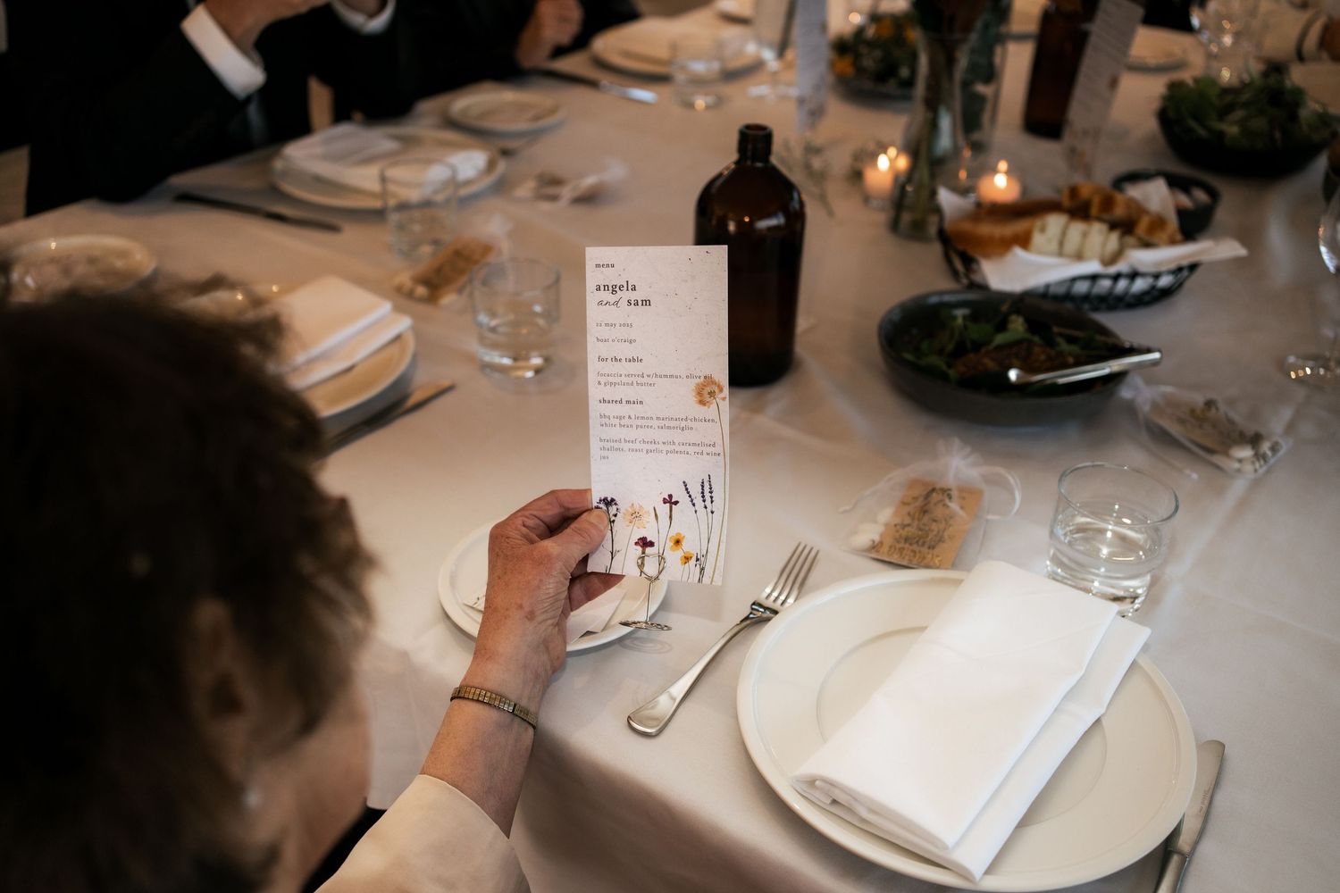Wedding guest reads menu card at elegant table setting with wine bottle and place settings.