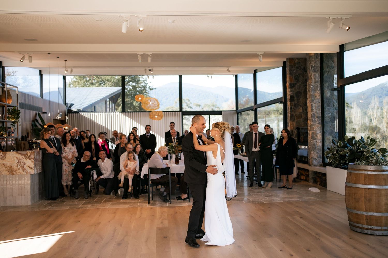 A couple shares their first dance in an elegant mountain venue with large windows and guests watching.