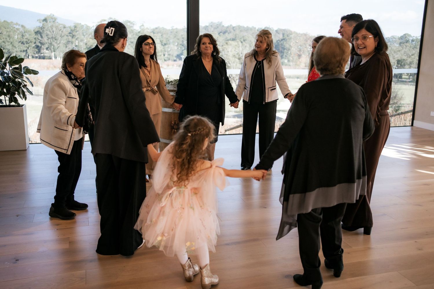 Wedding reception guests gather on a wooden dance floor with large windows overlooking scenic mountain views.