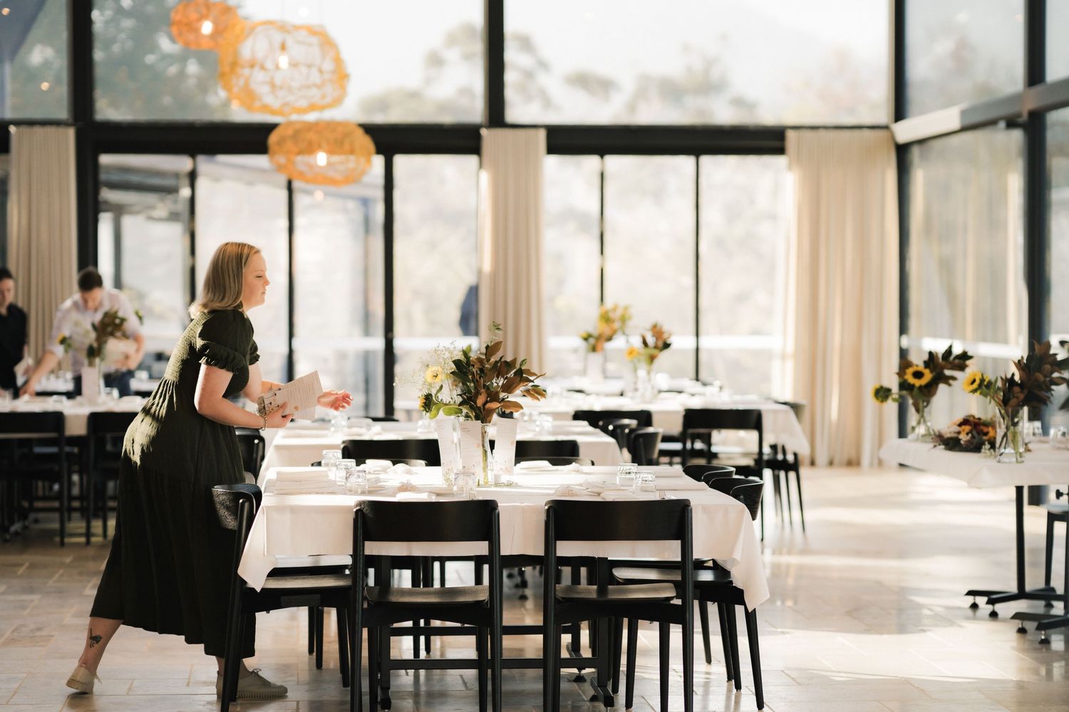 Event space with modern black chairs and white tablecloths being set up in a bright room with large windows.