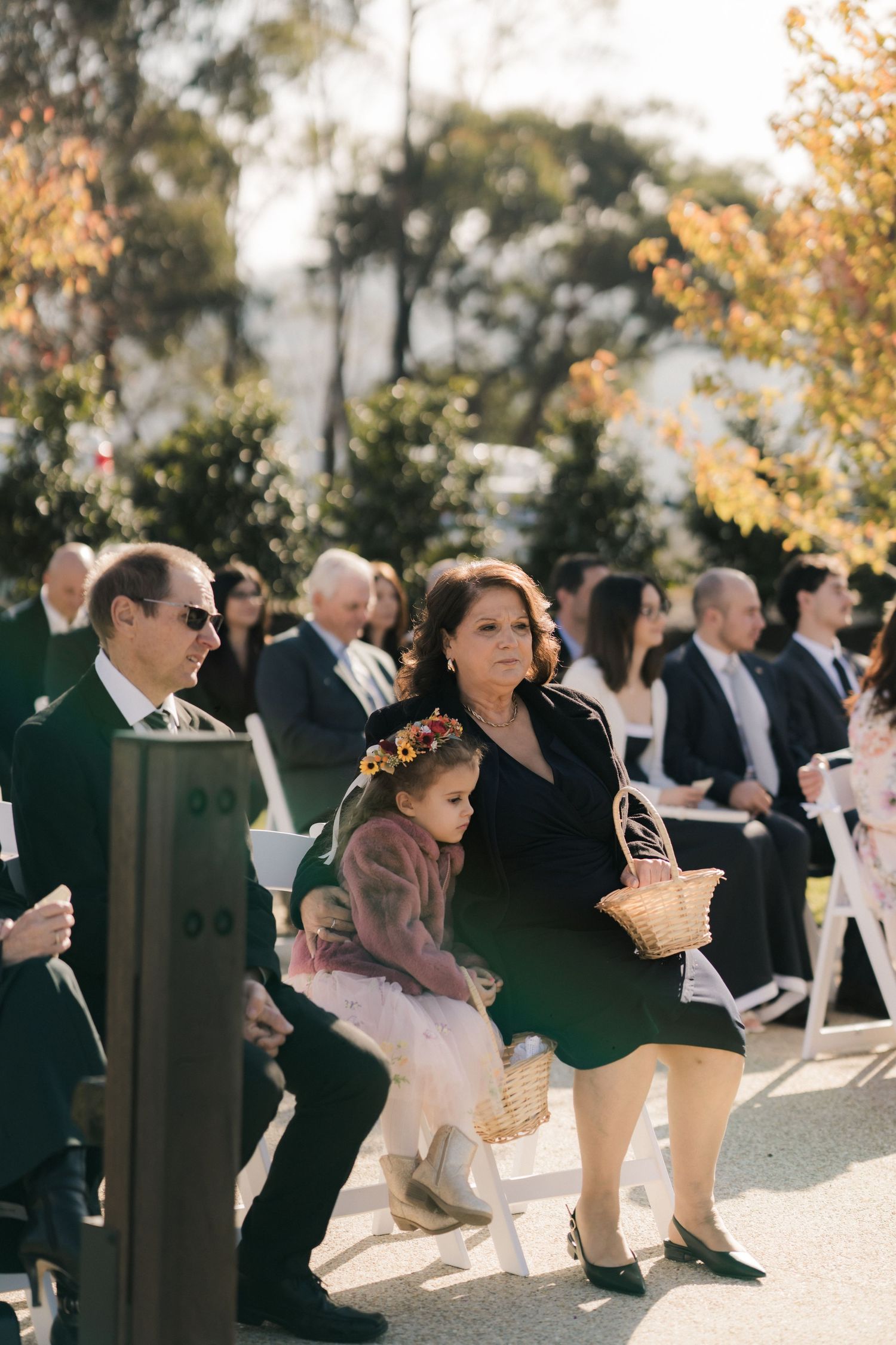Wedding guests sit outdoors in white chairs during an autumn ceremony beneath golden-leafed trees.