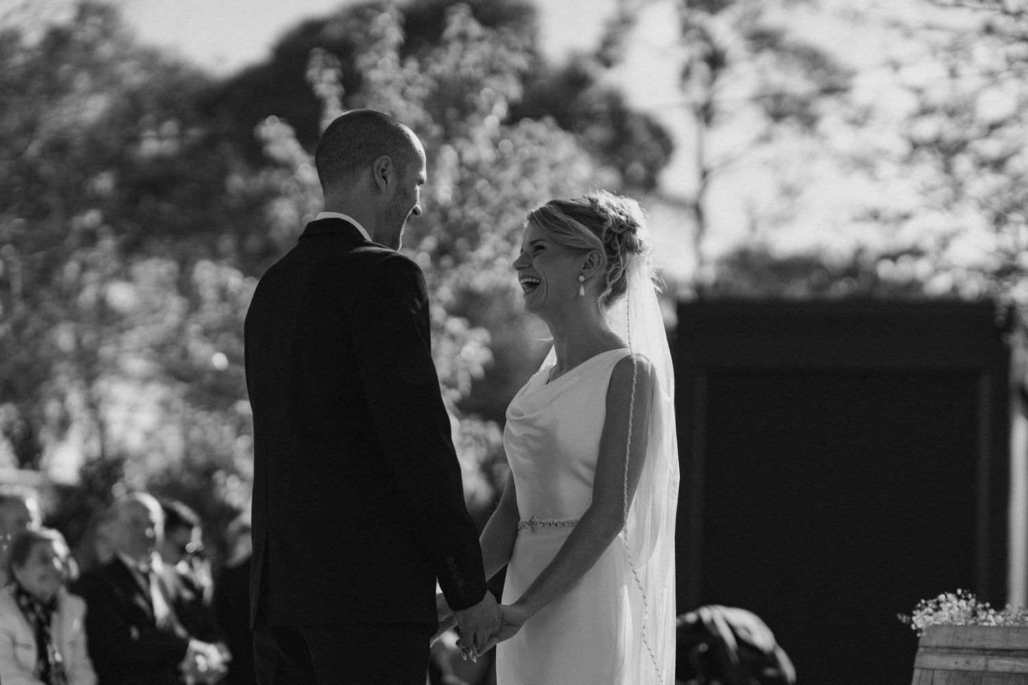 A romantic black and white wedding ceremony photo shows a couple holding hands while exchanging vows outdoors.