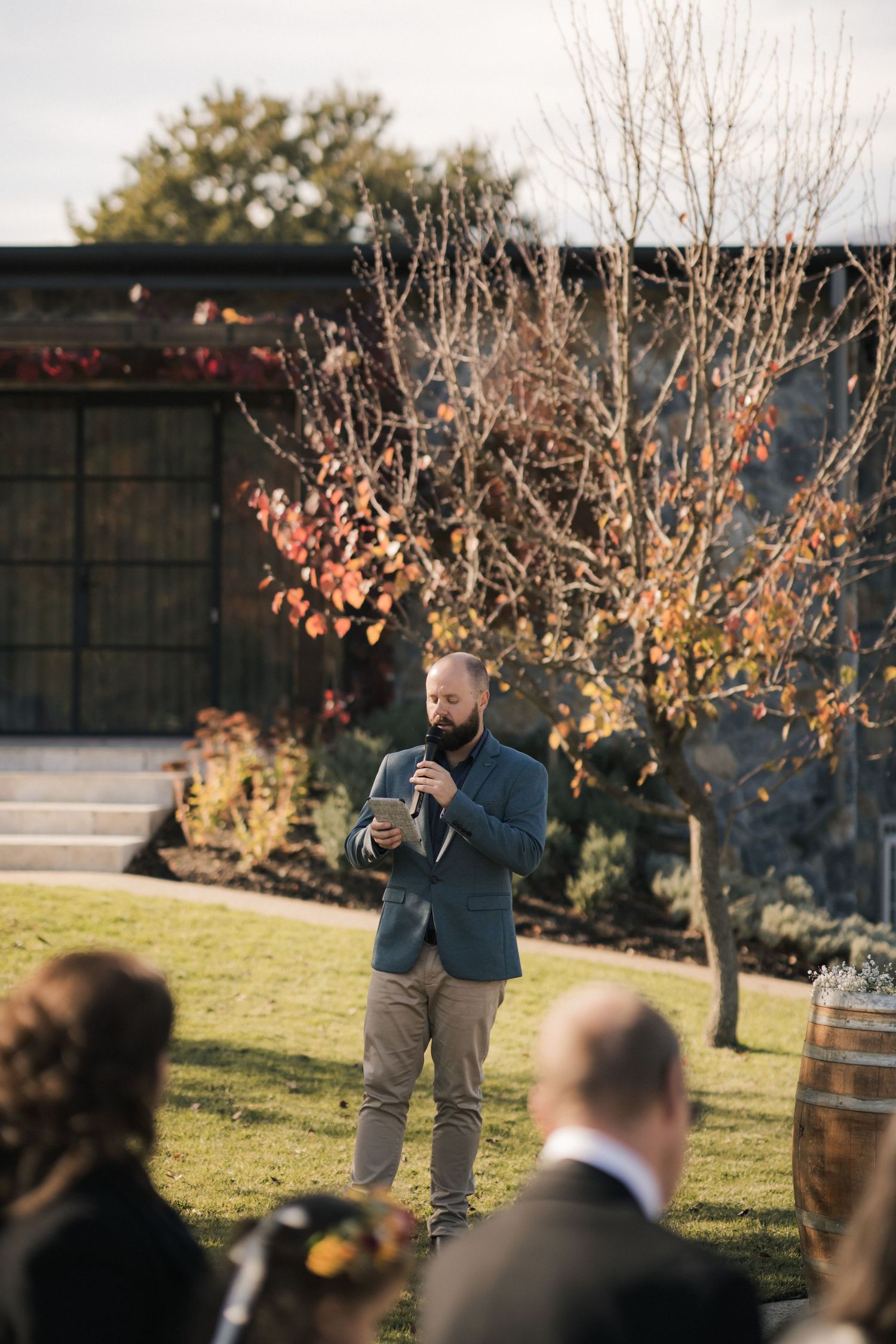 Someone speaking to guests during an outdoor fall wedding ceremony with autumn trees and foliage in the background.