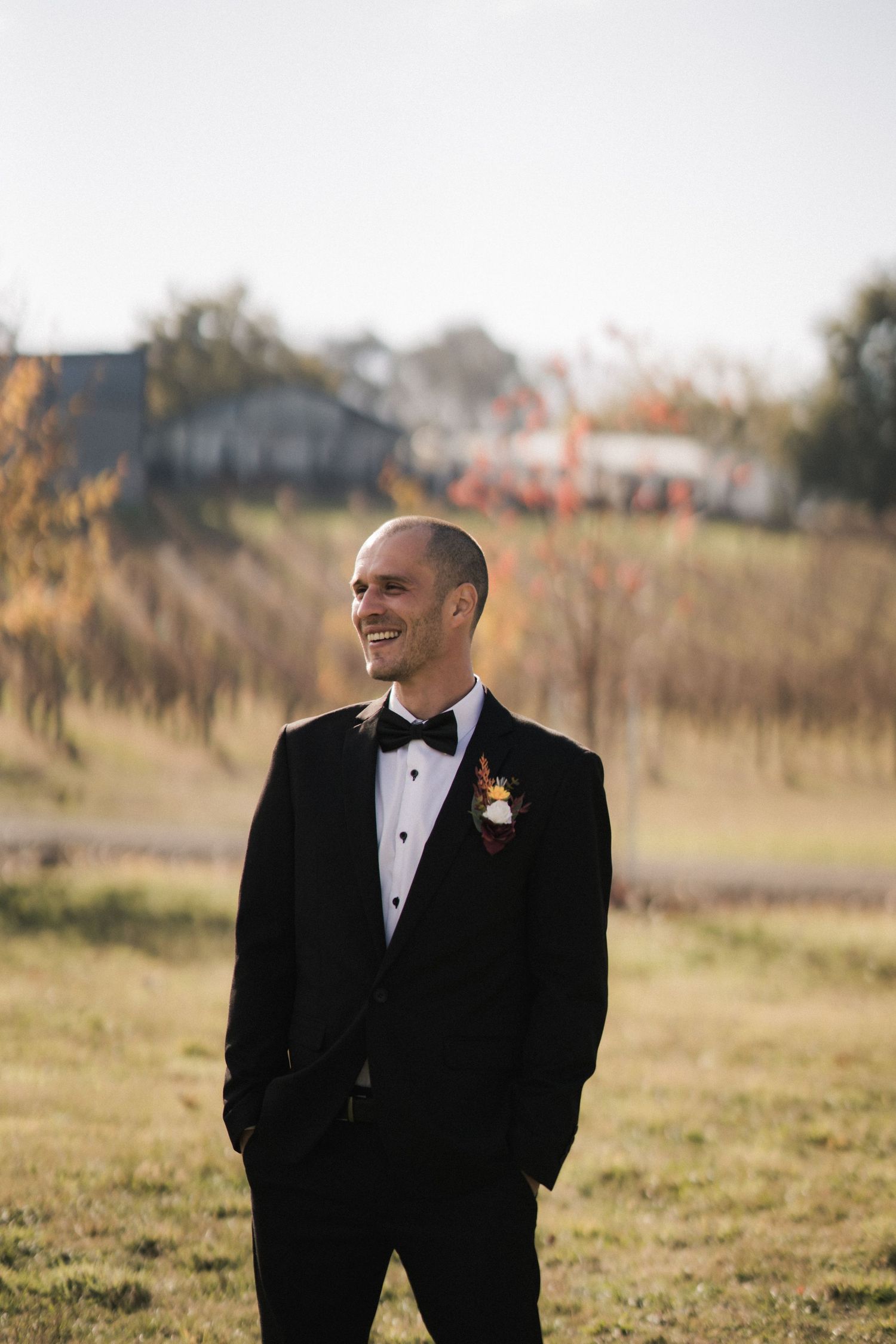 A groom in a black tuxedo stands outdoors in a grassy field during golden hour with houses in the background.