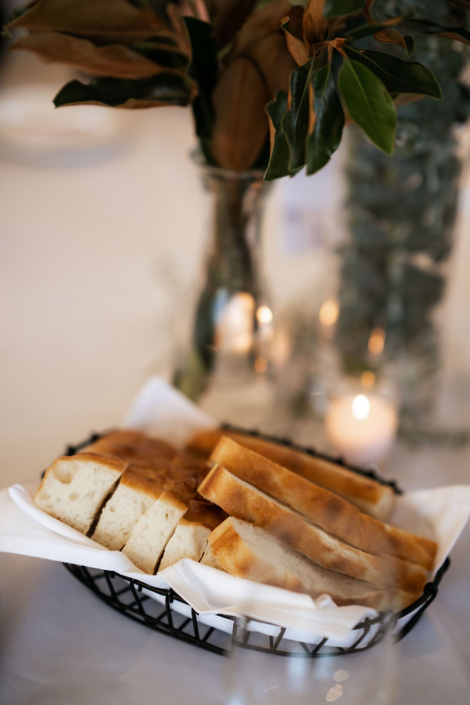 Sliced bread arranged in a wire basket with cloth lining, set against blurred candlelight and greenery in vases.