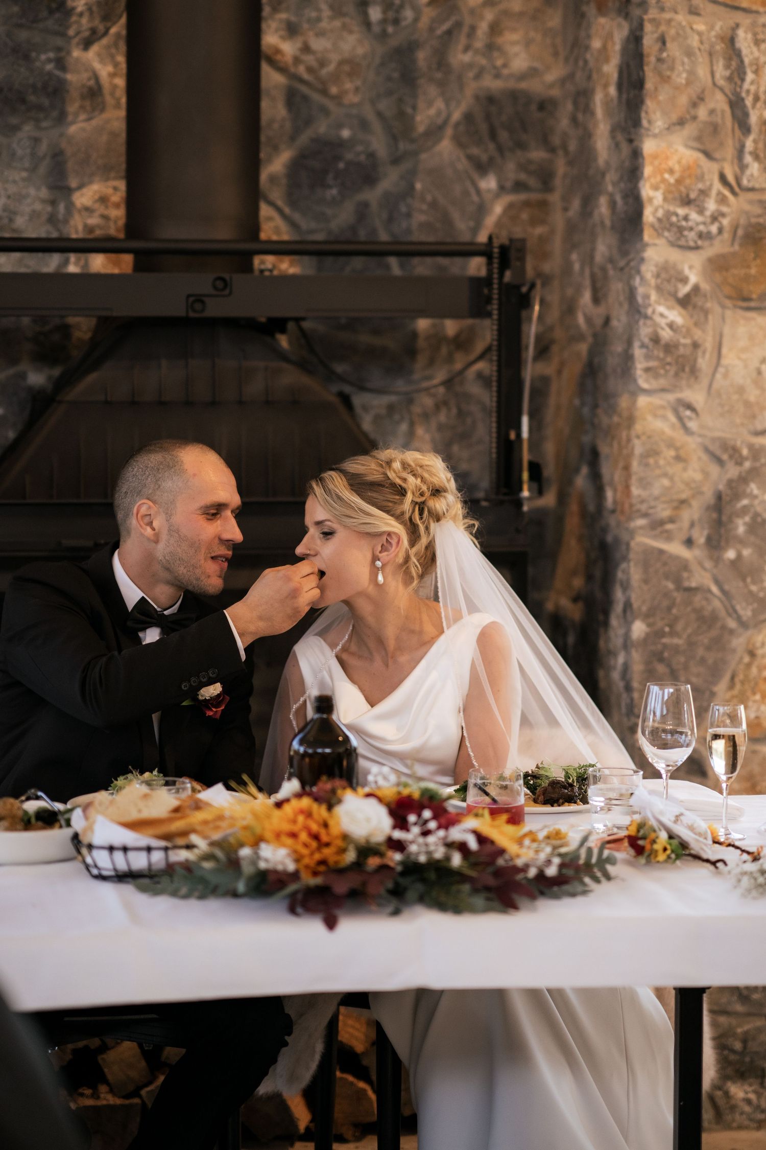 A couple shares an intimate moment during their wedding meal at a rustic stone venue with a decorated table.