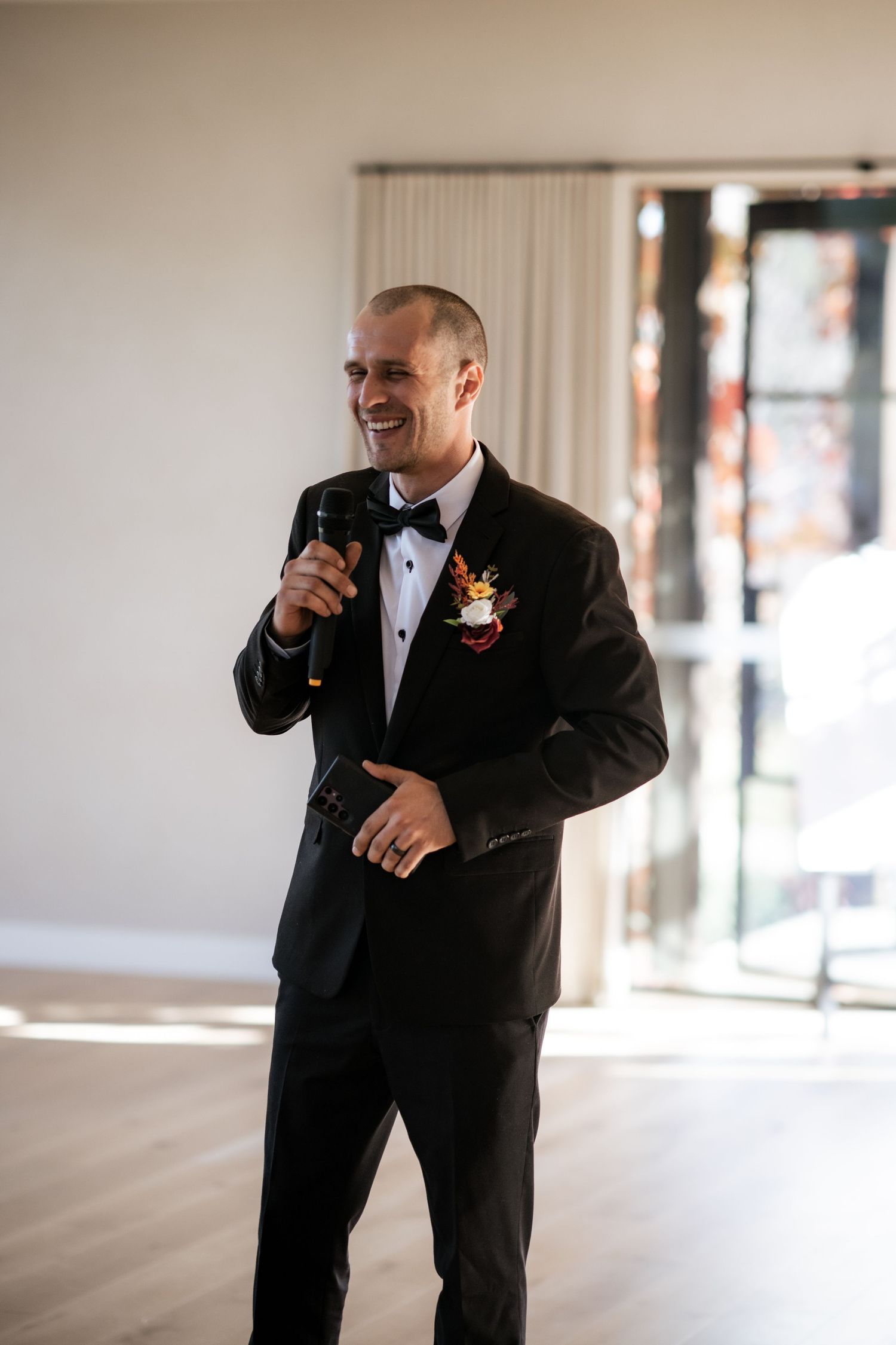 A groom in a black tuxedo giving a speech at his wedding reception.