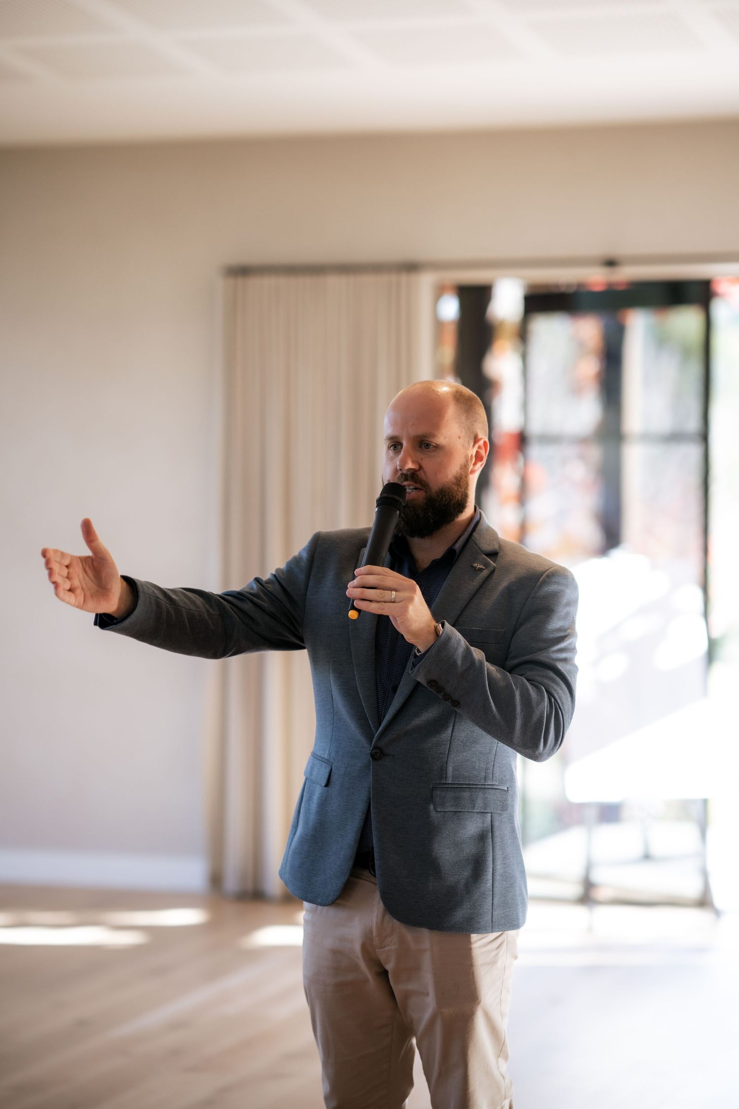 A person in a gray blazer speaks into a microphone while gesturing with their hand during a presentation.