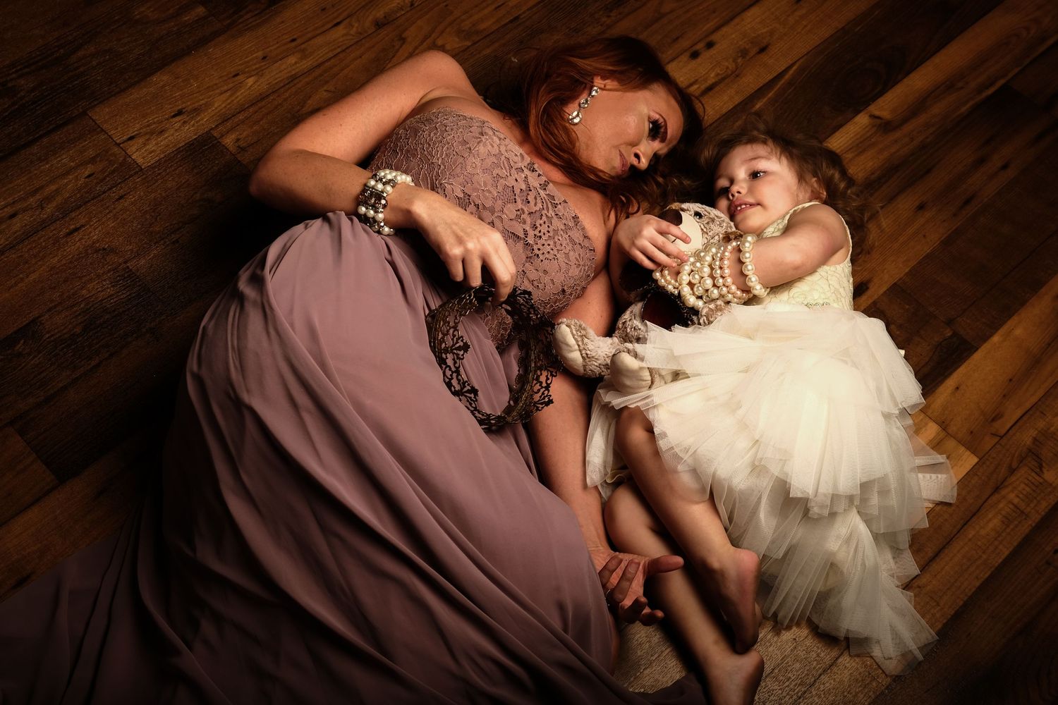 A woman in a mauve dress lying on wooden floor with a toddler in a white dress. Family Photography