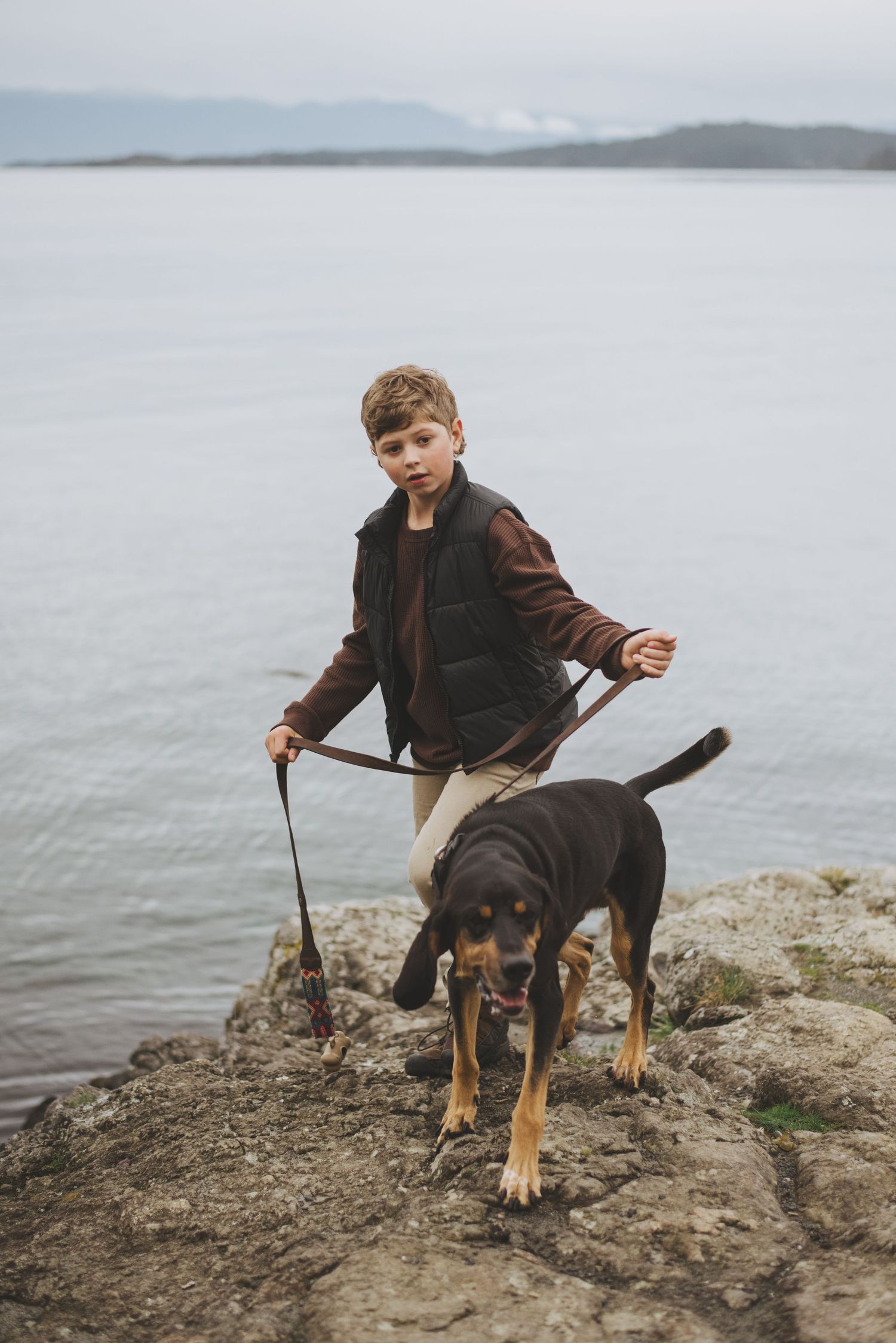 A young boy and his dog stand on a rocky outcrop overlooking the ocean with mountains in the distance.