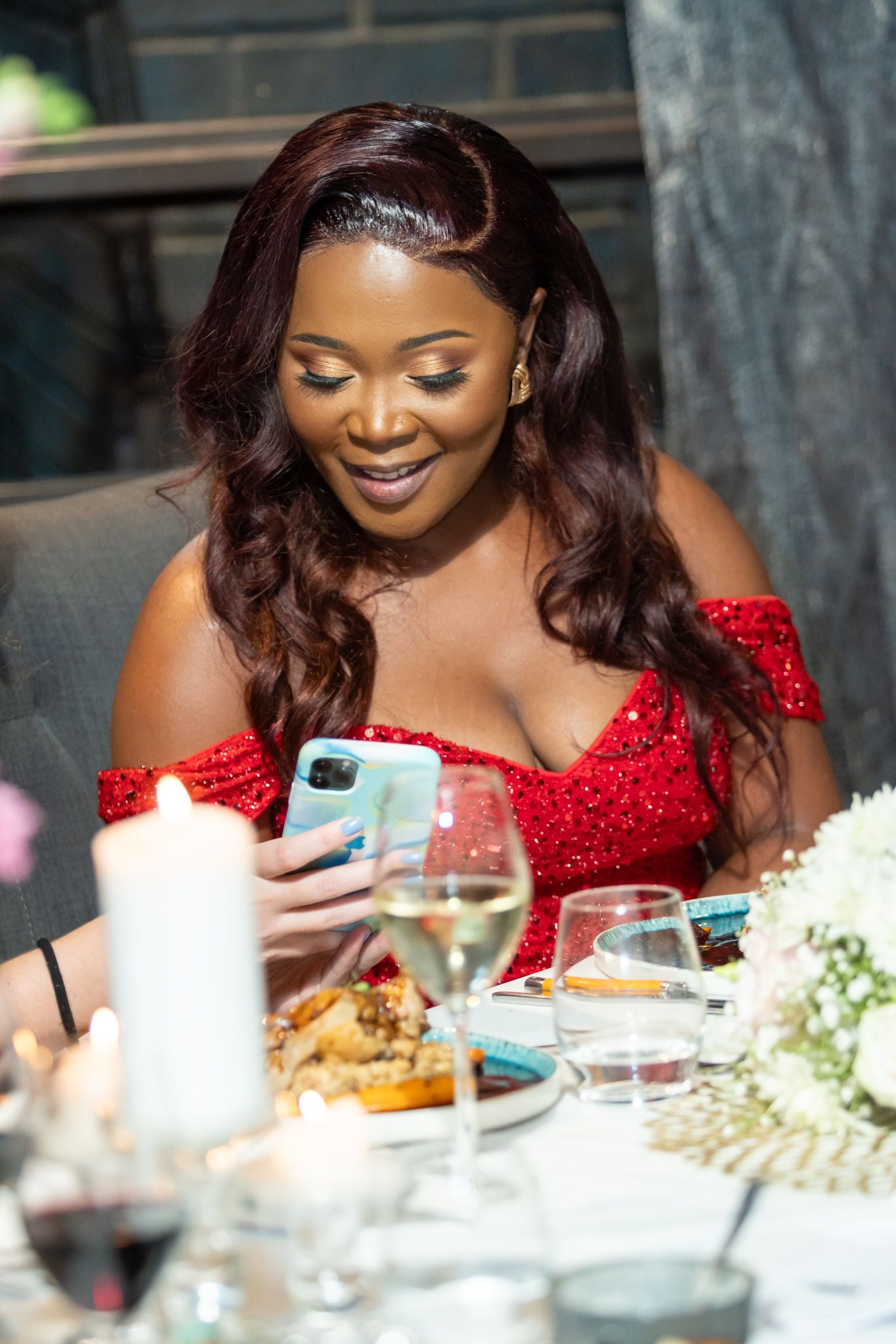 Woman in red off-shoulder dress smiles while looking at phone during elegant birthday dinner with family and friends.