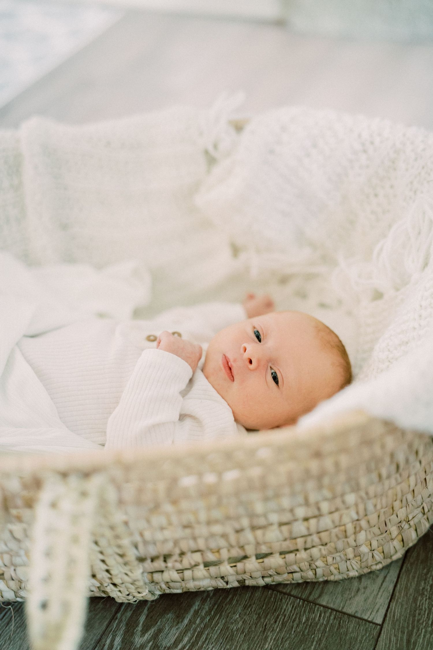 Baby in white outfit lying in woven basket on wooden floor.