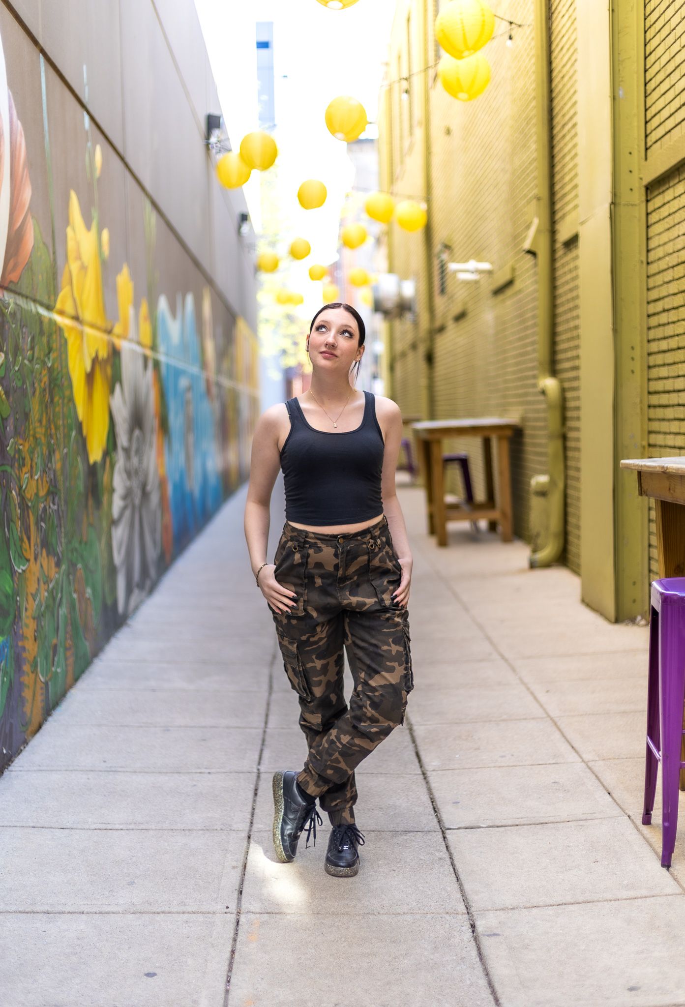 Urban alleyway decorated with yellow lanterns overhead, street art on walls and someone wearing camo pants and black top.