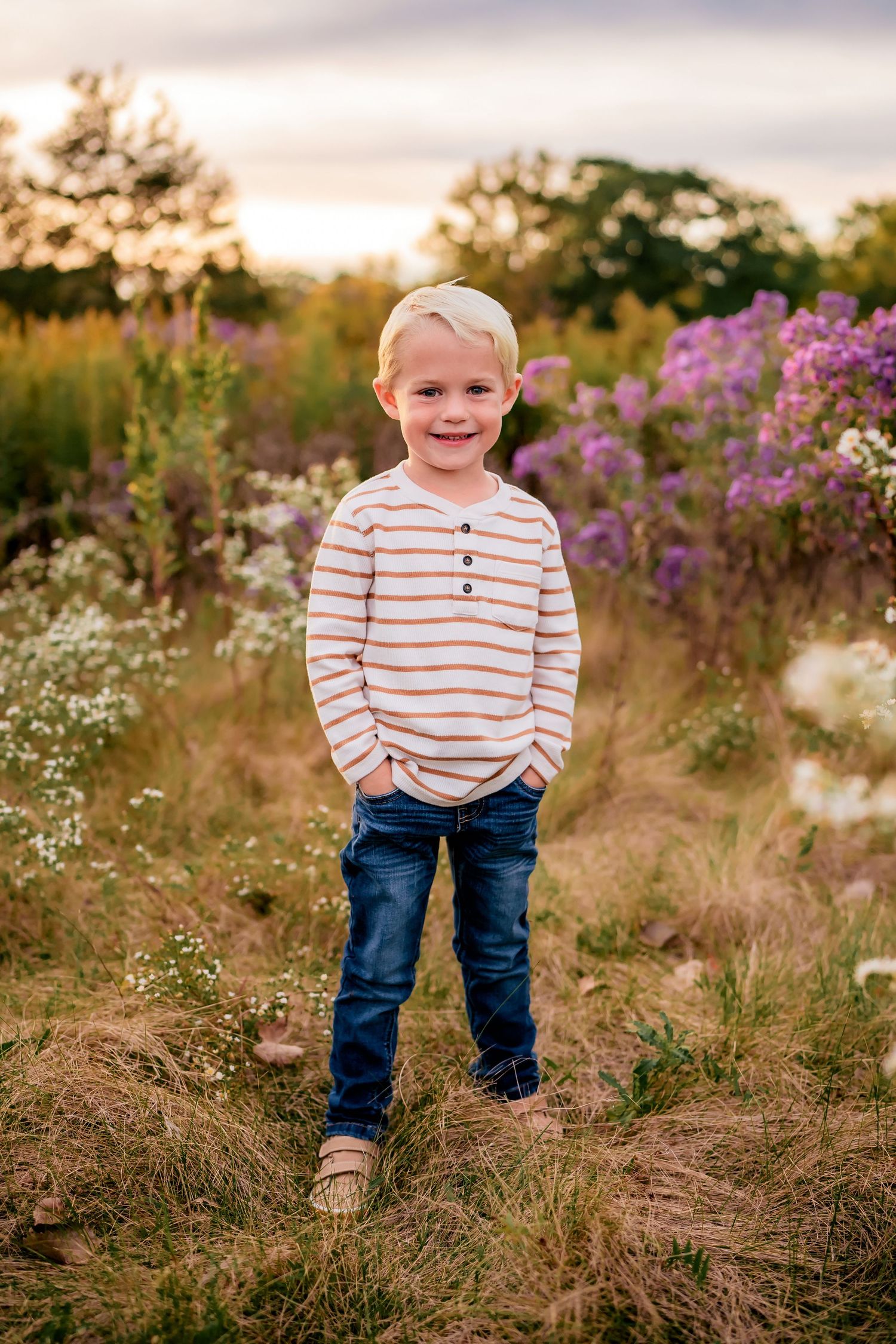 Young child in striped shirt poses in field with purple wildflowers at sunset.