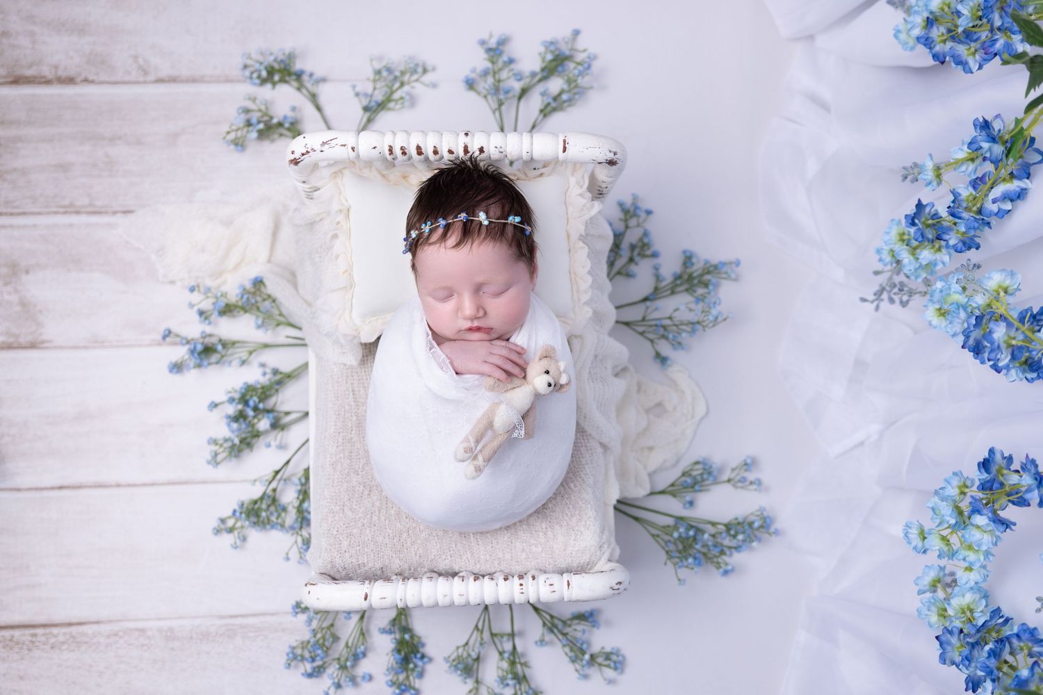 Sleeping newborn in white blanket nestled in white wooden box surrounded by blue and green floral decor.