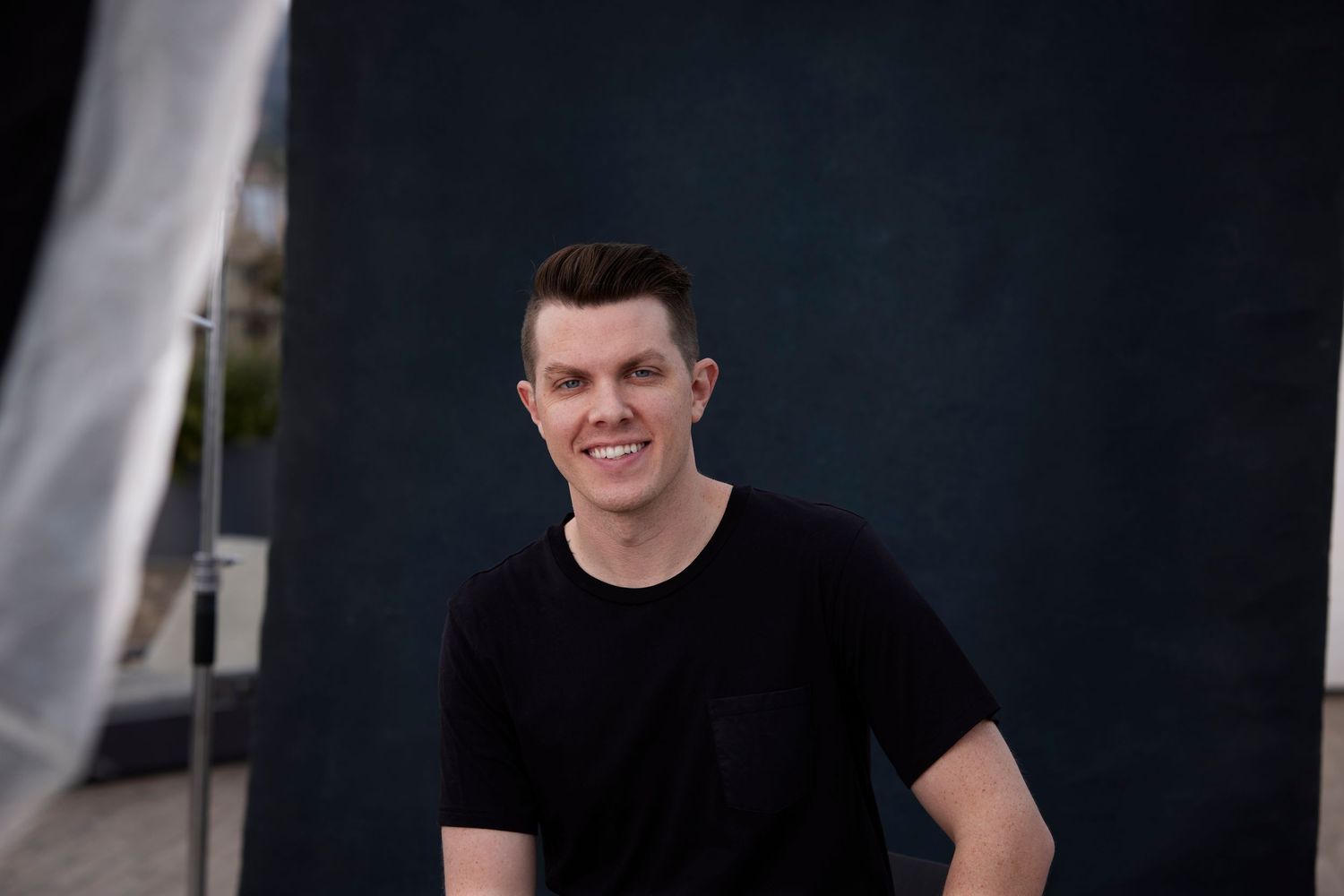 Studio portrait of photographer Adam Wamsley in black t-shirt smiling against dark painted background.